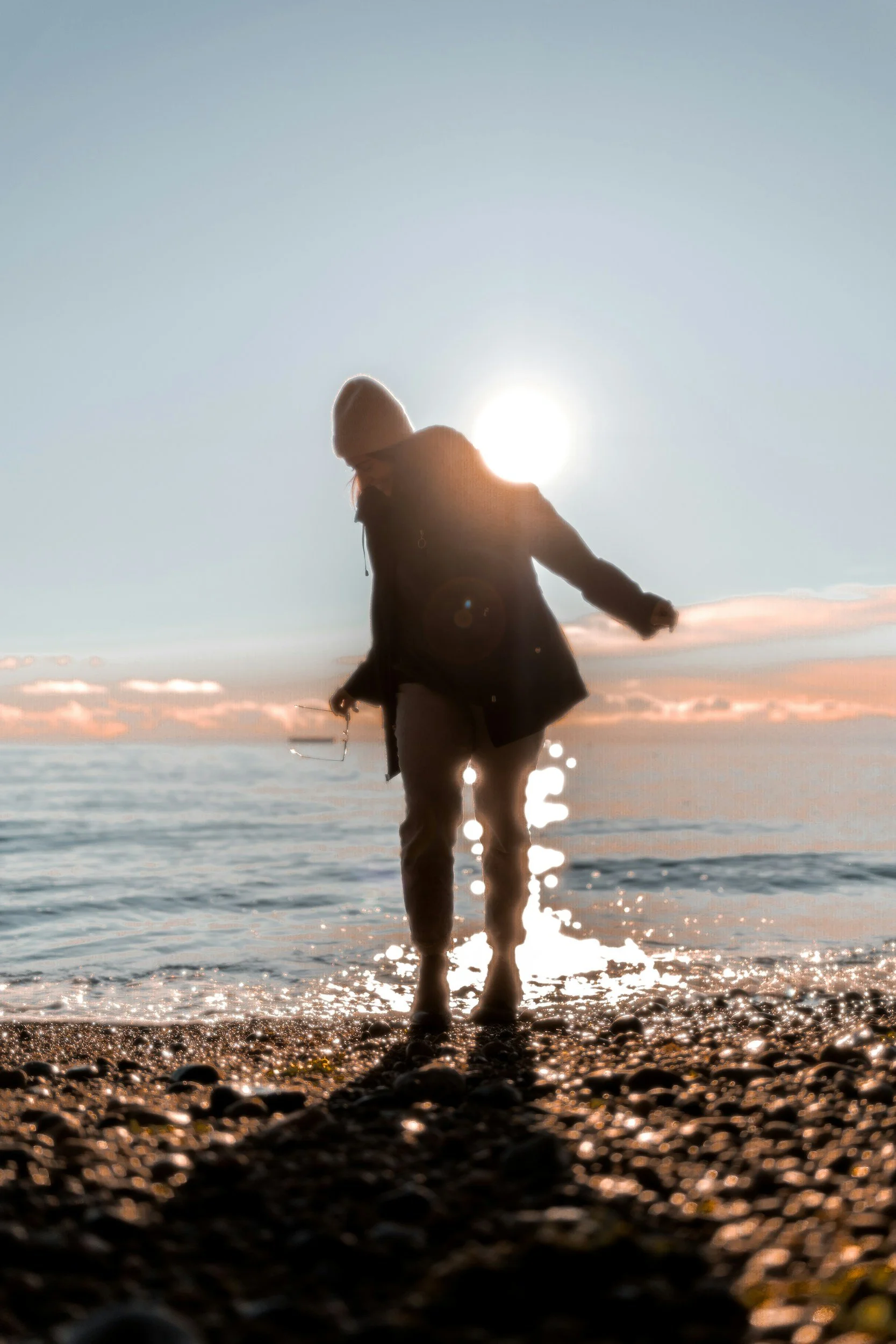 Person standing on a rocky beach at sunset, silhouetted against the sun with the sea in the background.