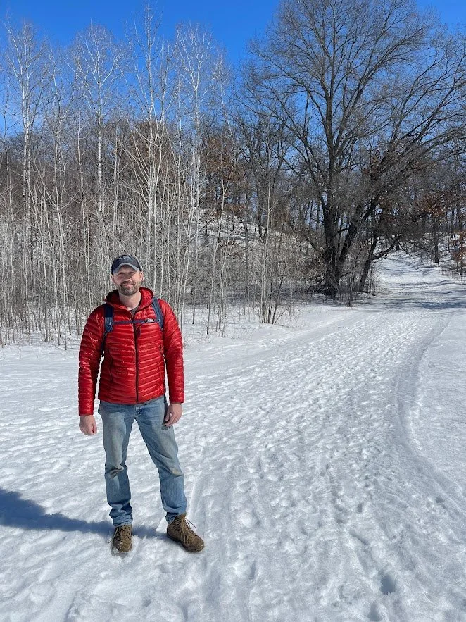 A man standing on a snowy trail in a winter landscape, wearing a red jacket, blue jeans, hiking boots, and a backpack, with leafless trees and a clear blue sky in the background.