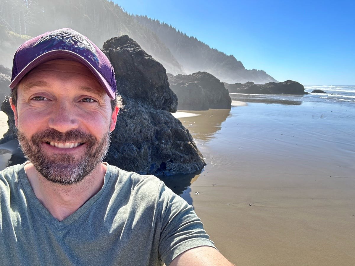 A man with a beard and a purple cap taking a selfie on a beach with large rocks and cliffs in the background, under clear blue skies.