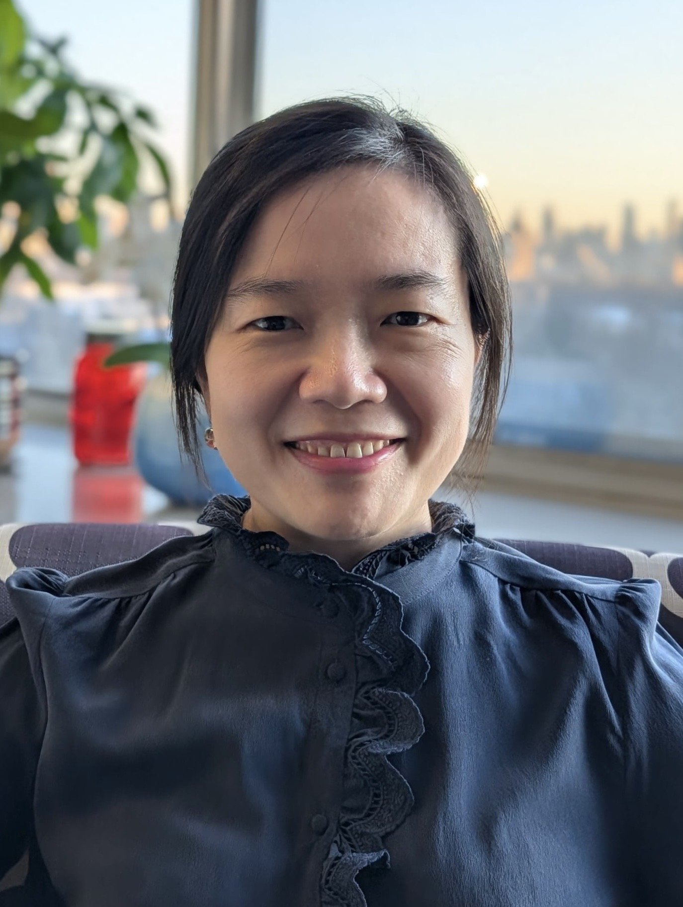 A woman smiling at the camera with a city view and sunset in the background, sitting indoors near a window.