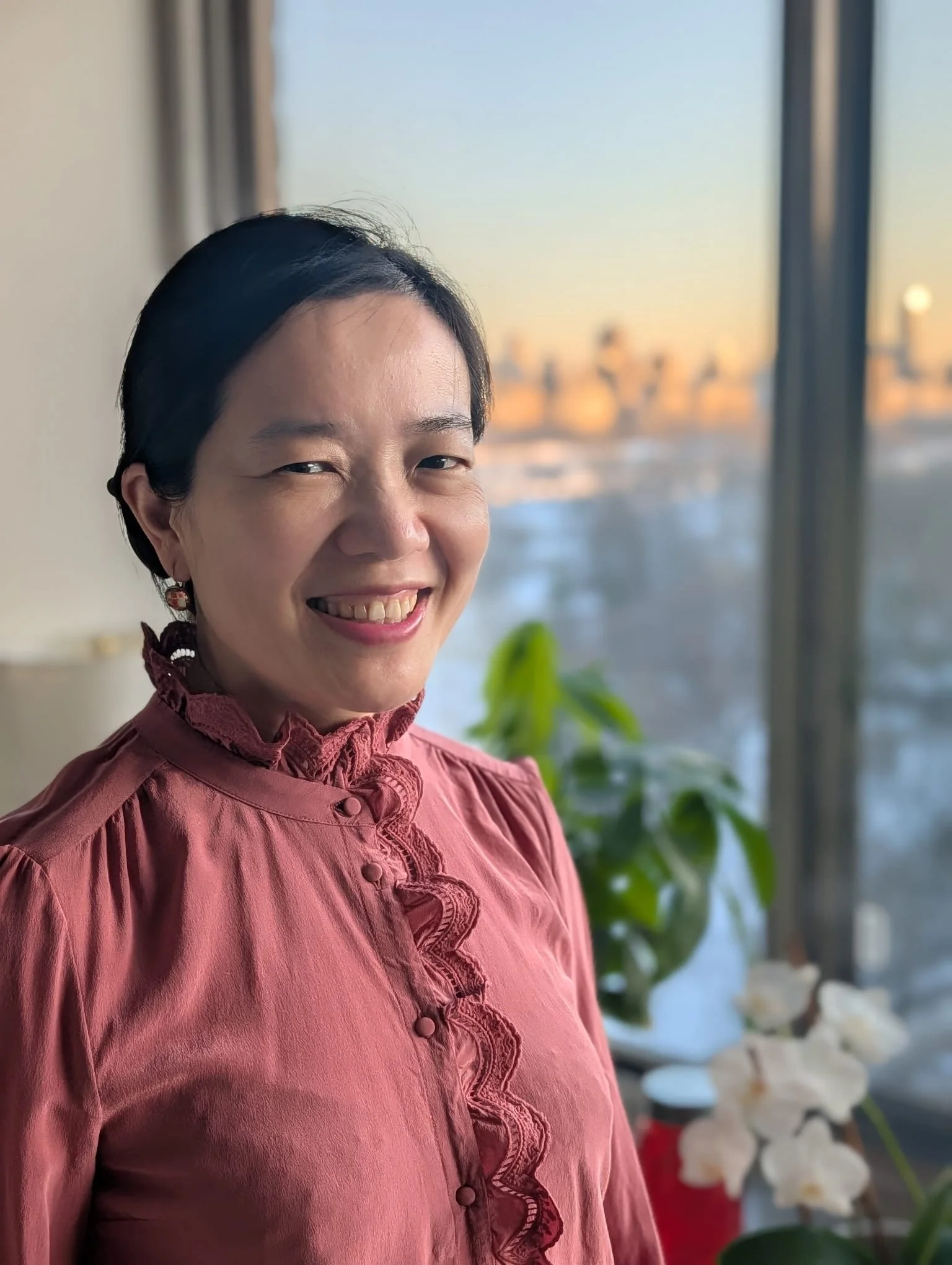 Smiling Asian woman with short black hair wearing a pink top with ruffles on the front, standing indoors near a large window with a cityscape in the background during sunset, with plants and flowers nearby.
