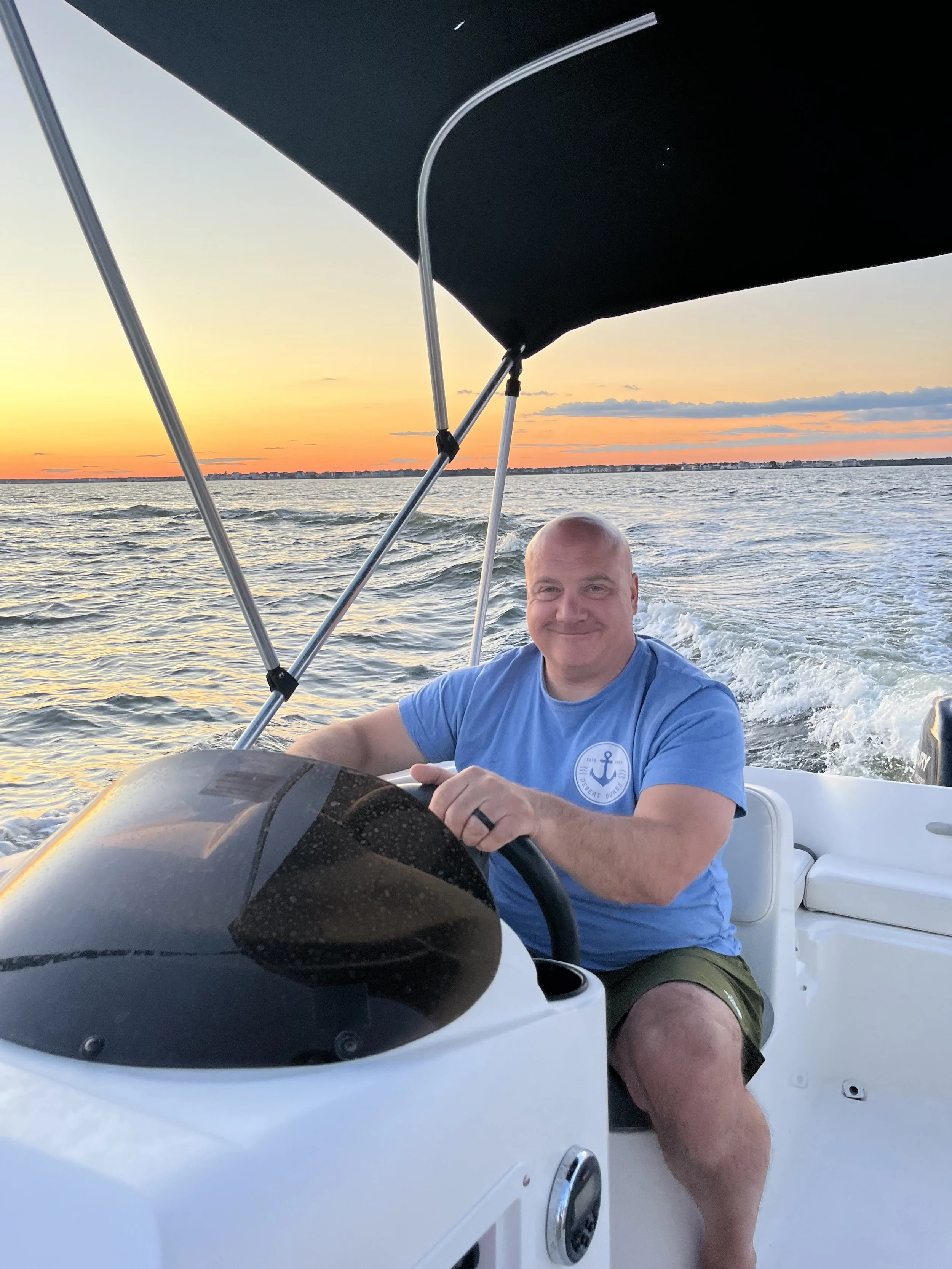 Man with a bald head smiling while steering a boat on the water at sunset, wearing a blue shirt with an anchor logo.