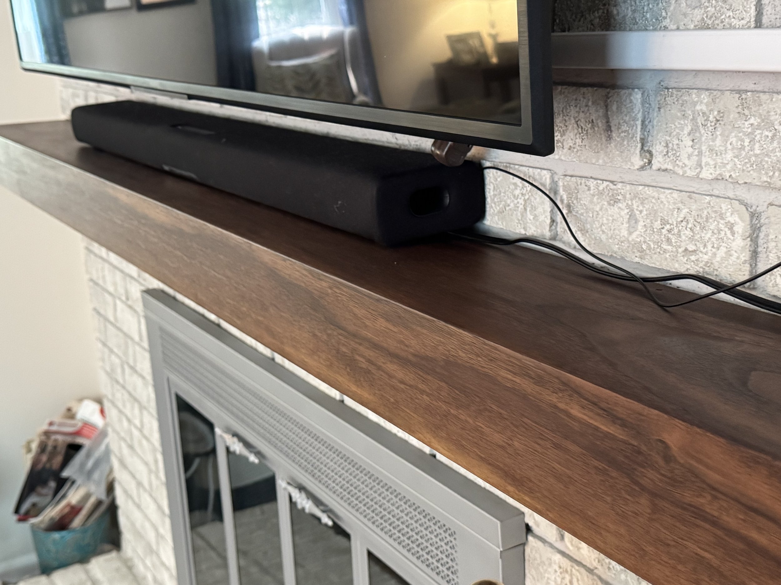 A television on a wooden mantel with a soundbar placed underneath it, against a white brick wall.
