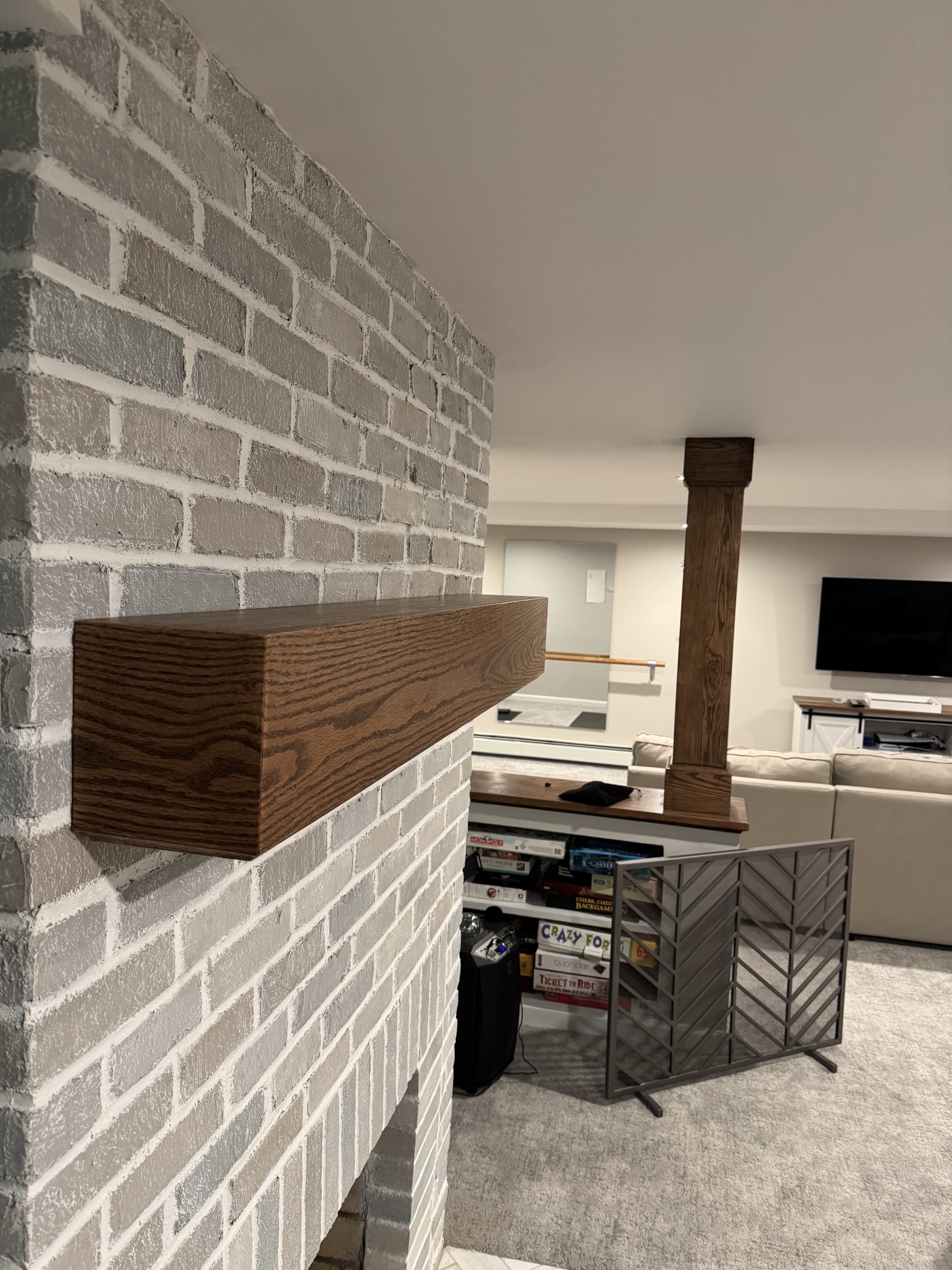 Close-up of a white brick fireplace with a wooden mantel on wall, overlooking a living room with a beige couch, a TV, and a game table.
