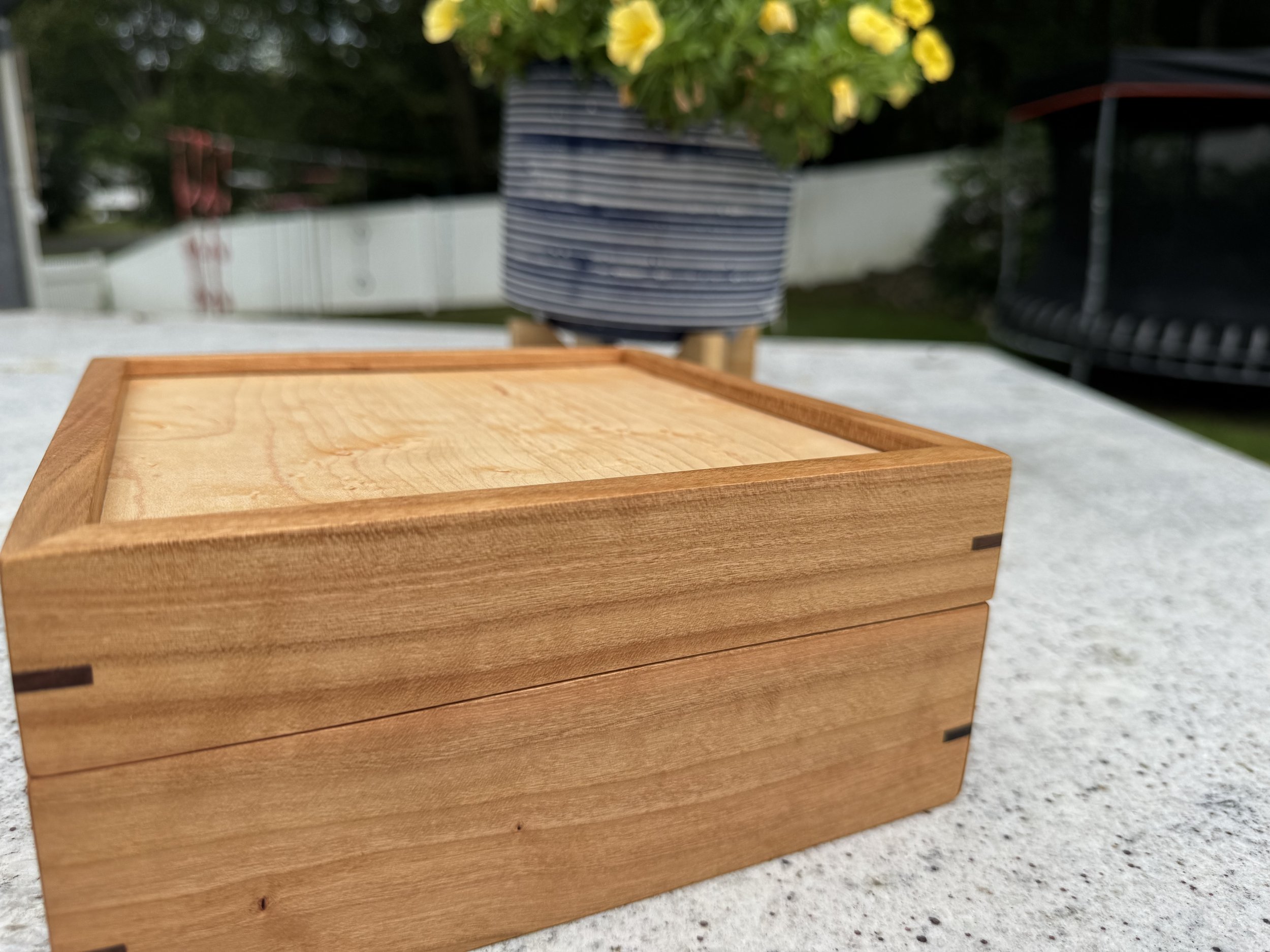 A wooden cherry box with dovetail joints placed on a stone surface outdoors, with a potted plant with yellow flowers, a white fence, and a trampoline in the background.