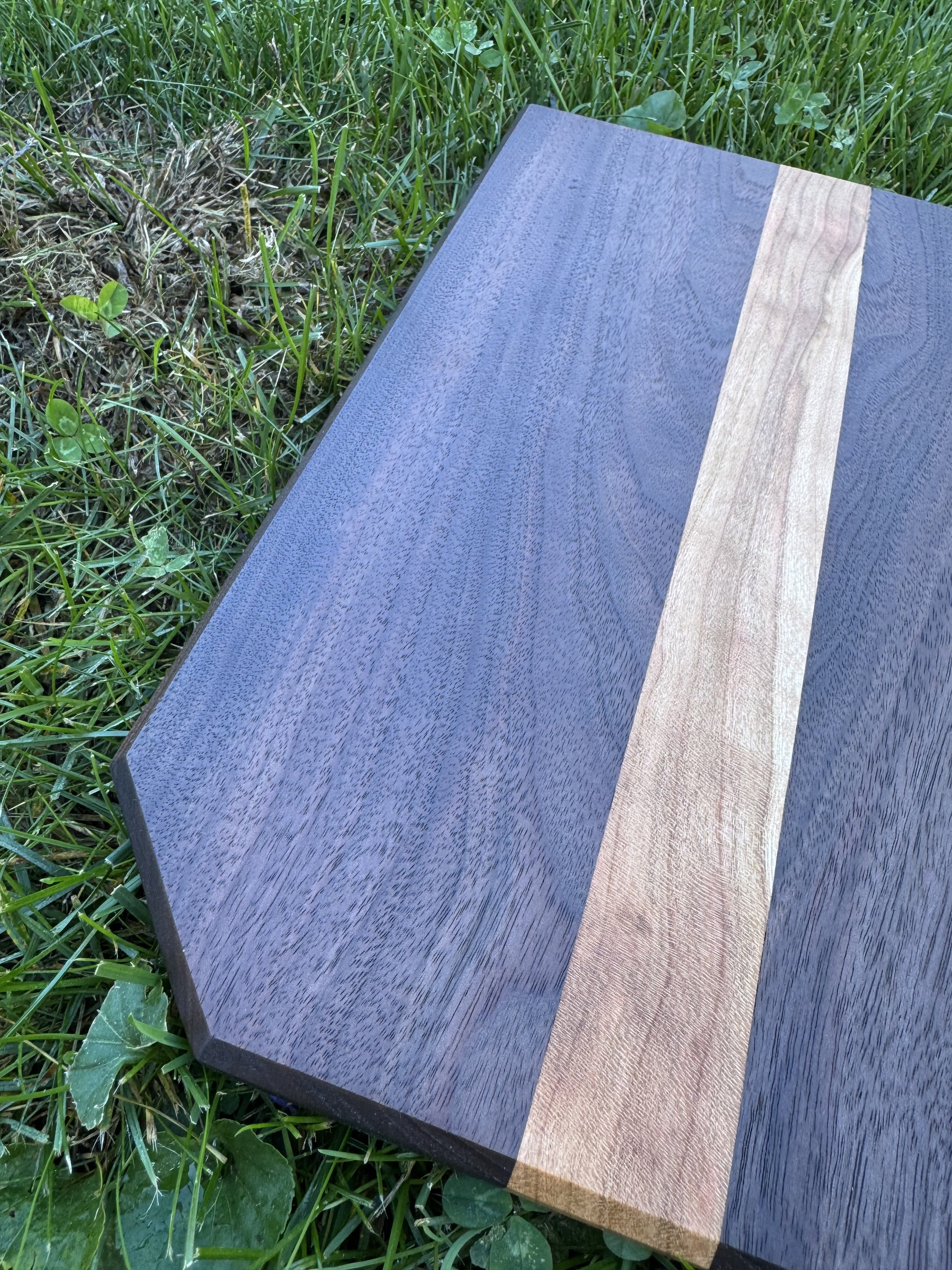 A wooden cutting board with a walnut wood section and a cherry wood strip, placed on green grass.