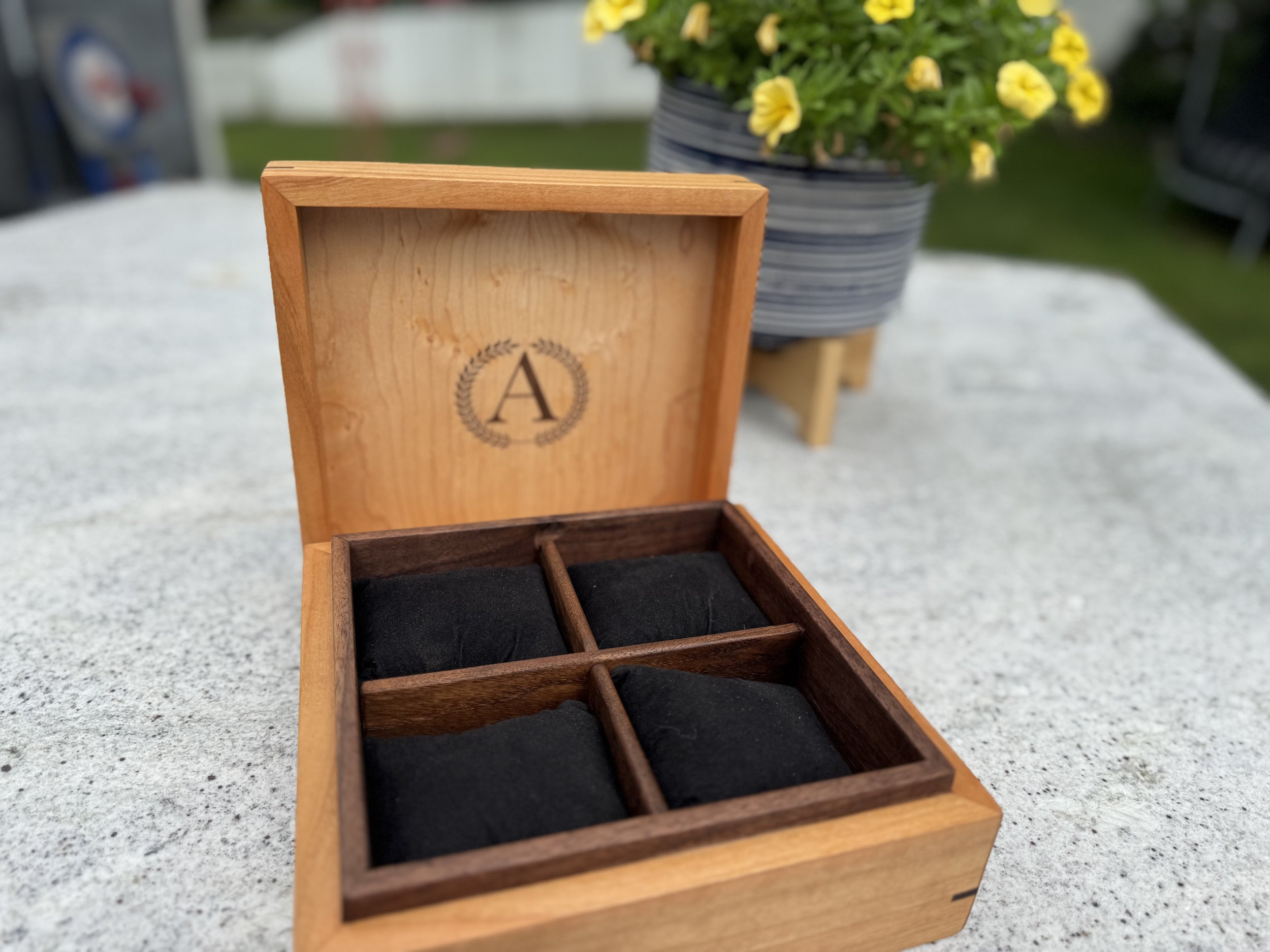 A wooden box with a monogram 'A' inside the lid and four compartments containing black cloth pockets, placed on a concrete surface outdoors.