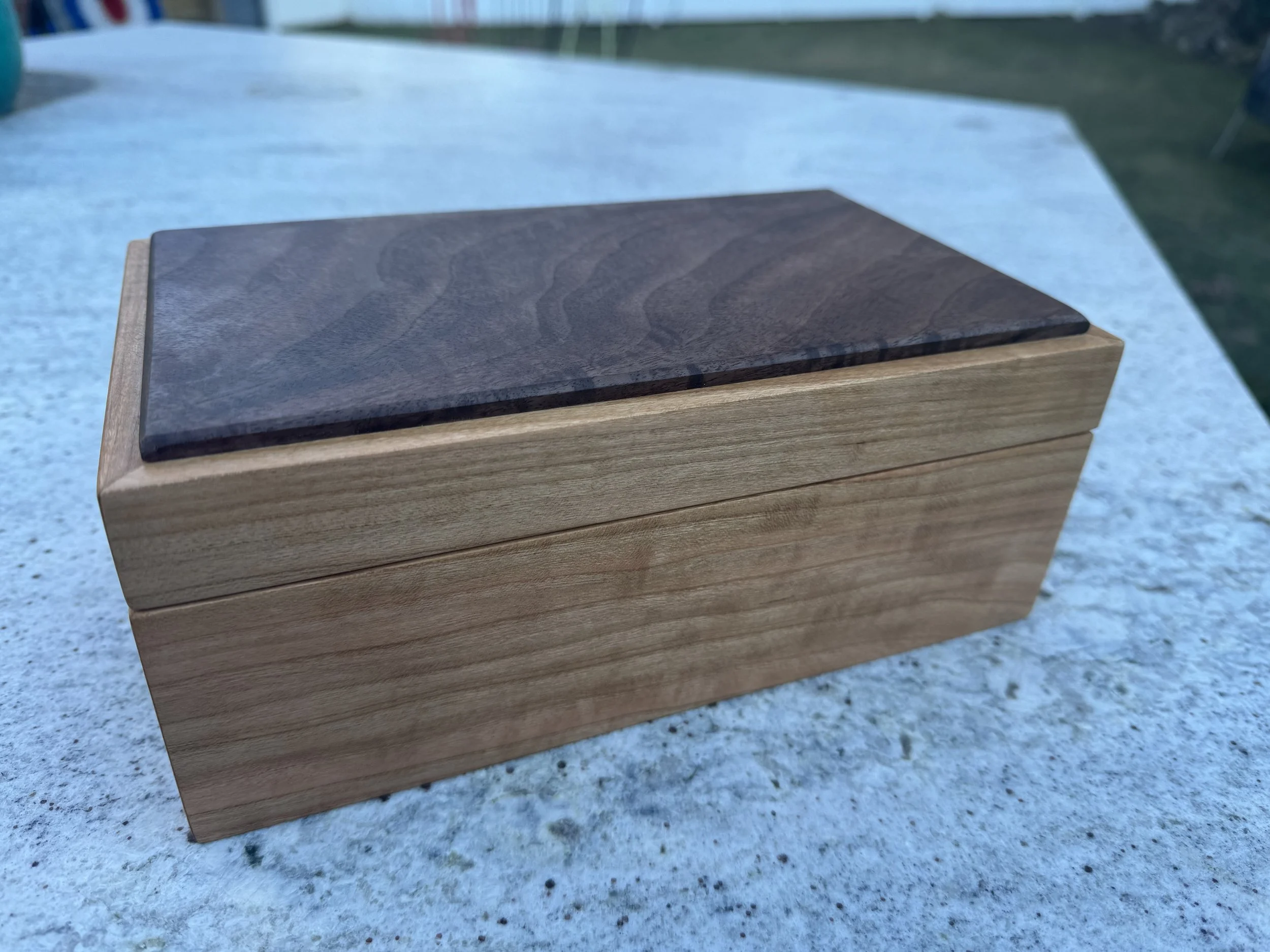 A rectangular wooden box with a dark wood lid on a light stone surface outdoors.