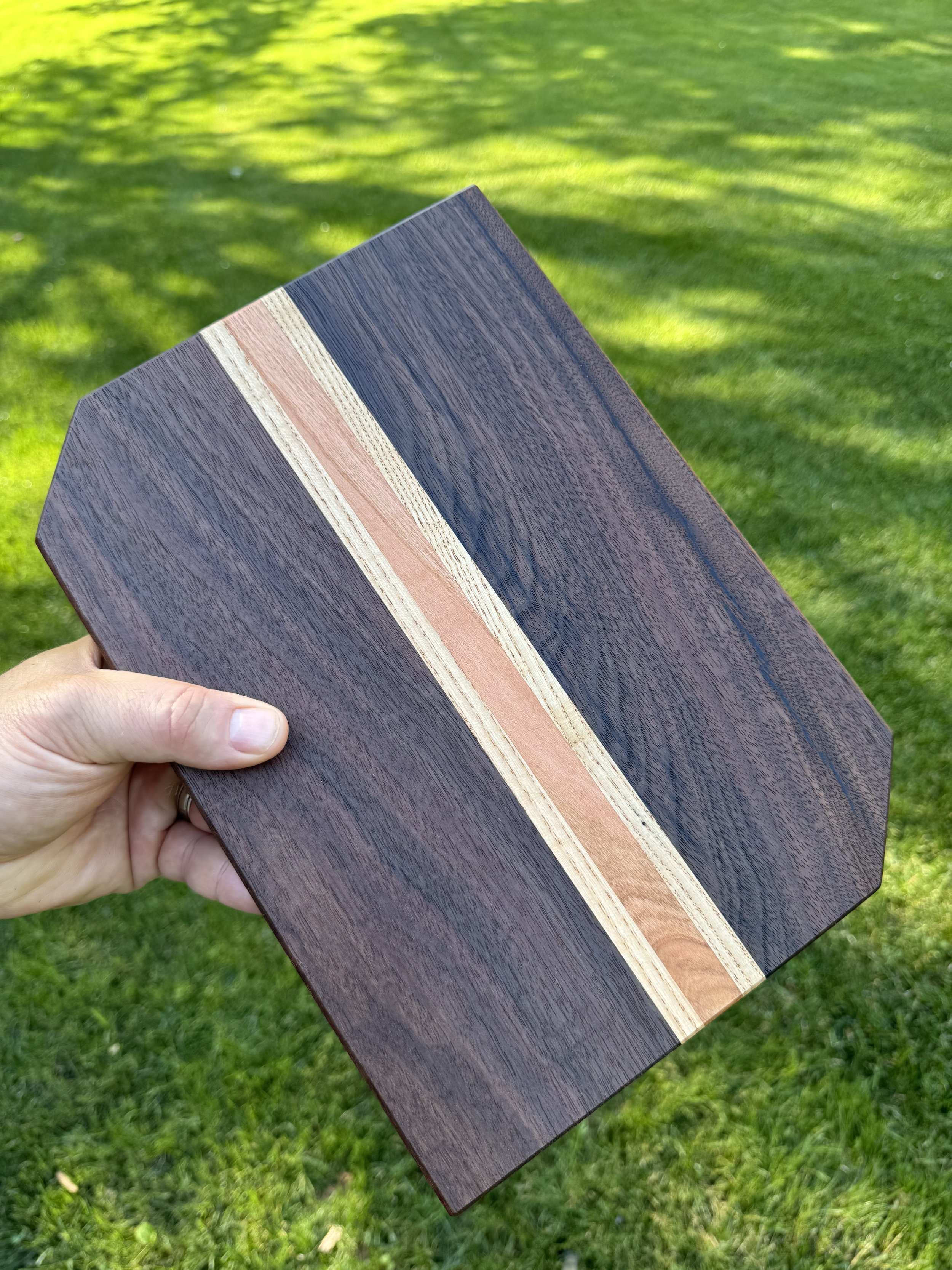 A hand holding a beautifully crafted wooden cutting board with a striped pattern in a grassy outdoor setting.