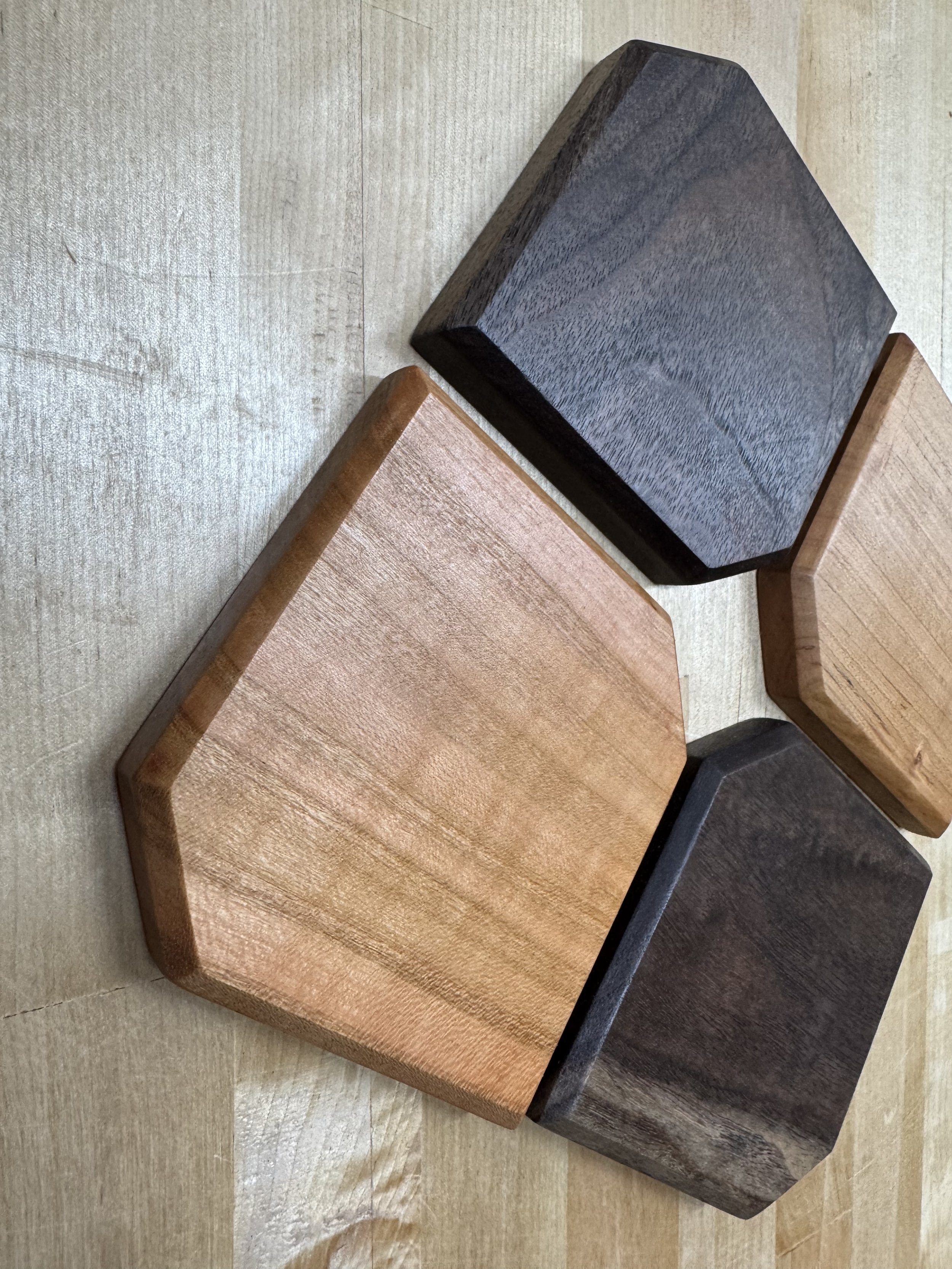 Four wooden hexagonal blocks in natural, dark, light, and black wood finishes, placed on a light wooden surface.