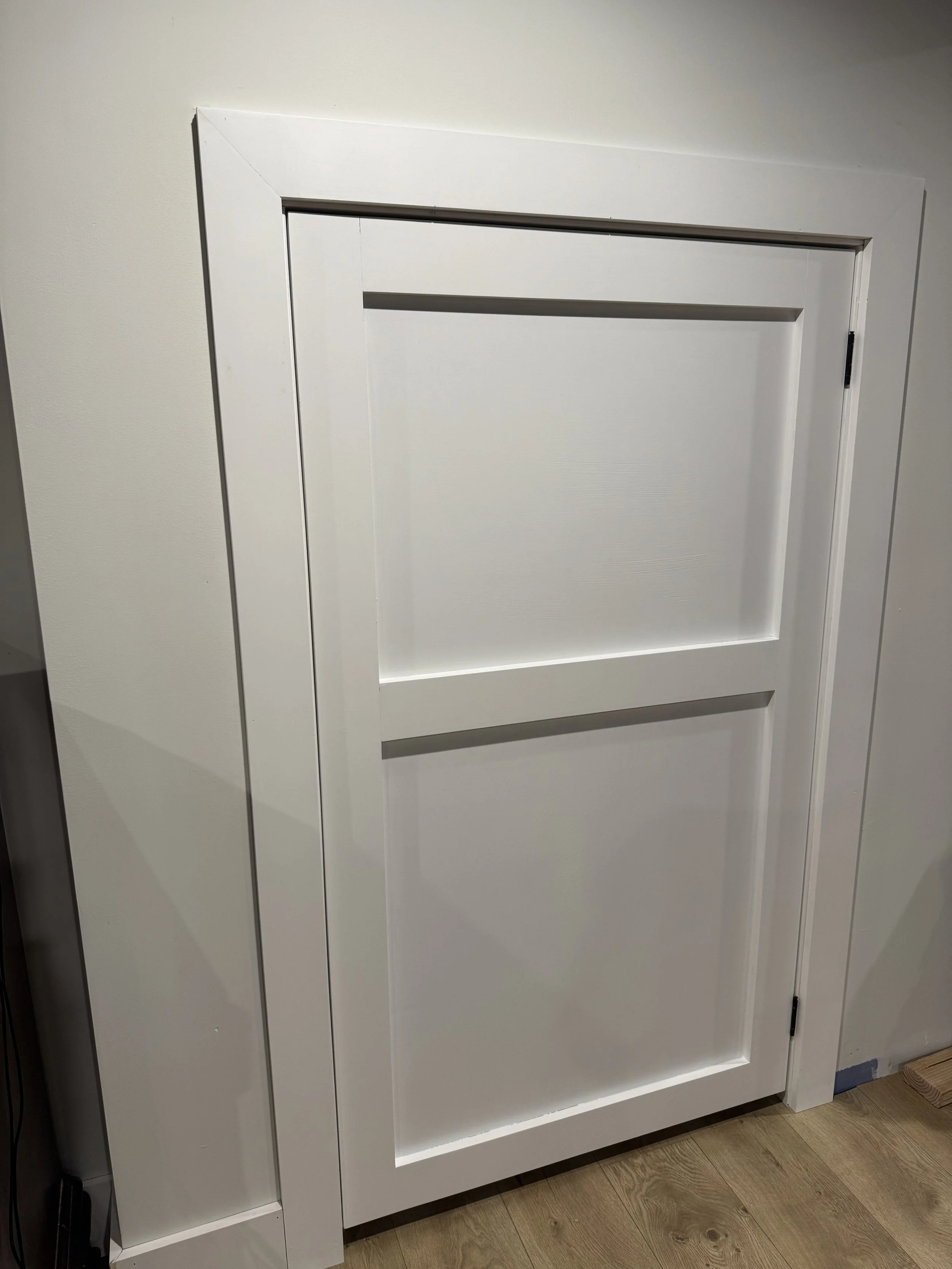 Close-up of a white interior door with a simple panel design, mounted on a white wall with wooden flooring.