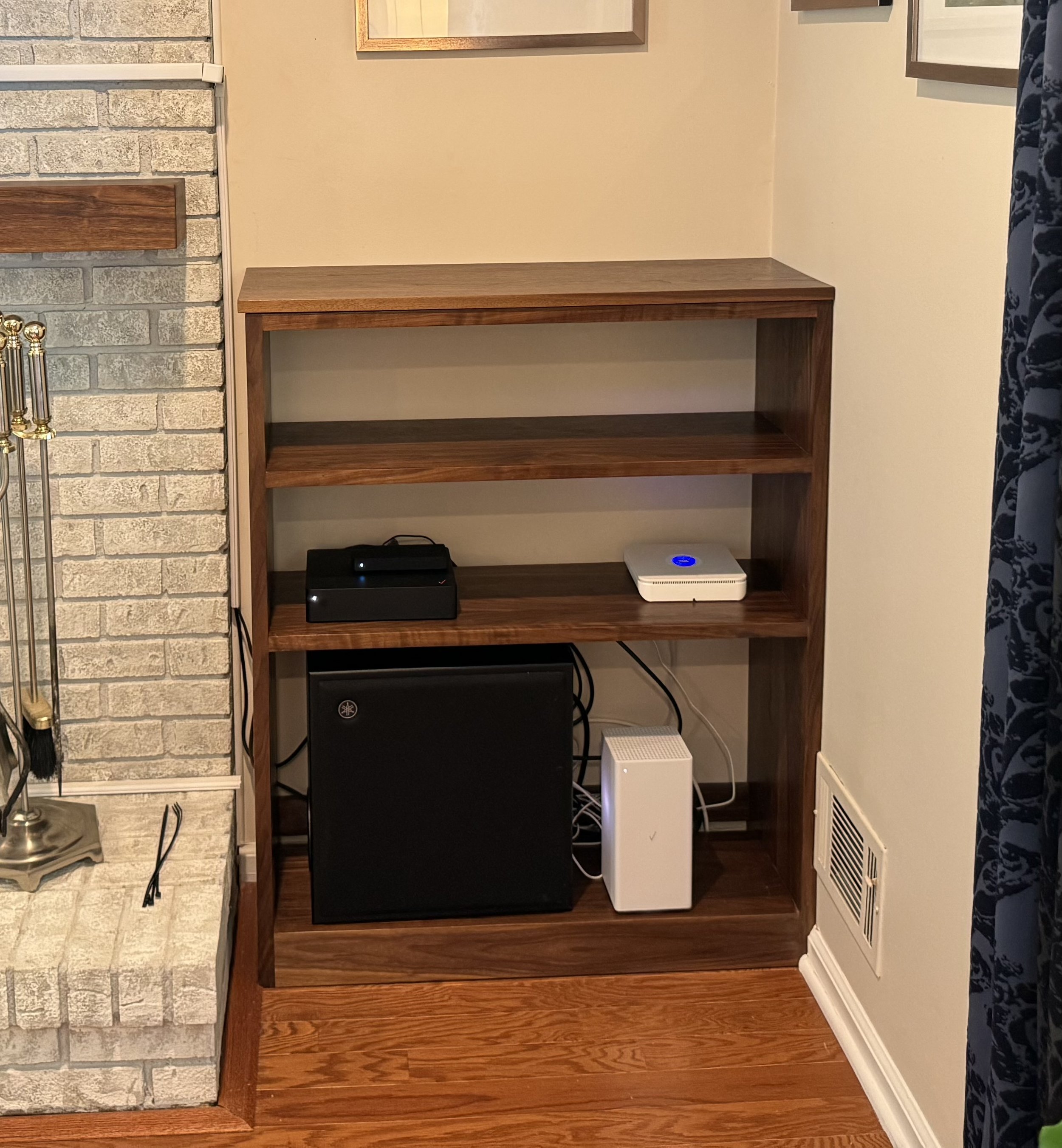 Wooden bookshelf containing a black subwoofer, a white device on the middle shelf, a black device on the middle shelf, and a white device on the bottom shelf, with cables connected to the devices.