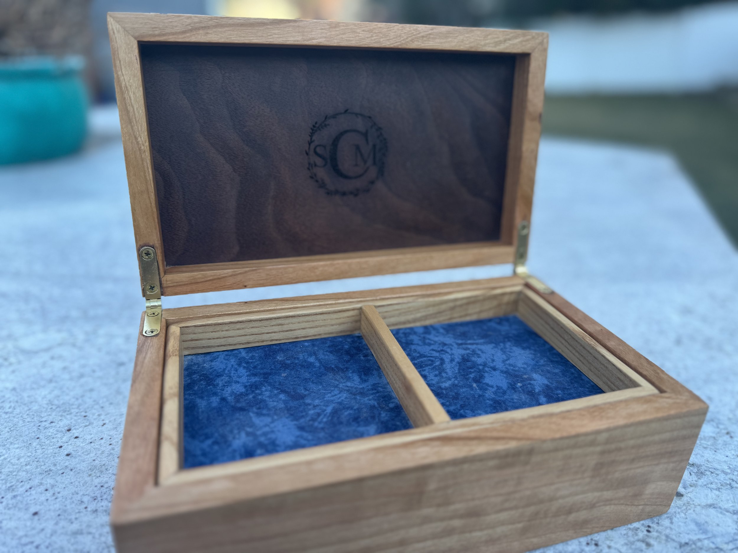 Open wooden box with divided blue velvet interior and a transparent glass lid with initials 'C' and 'M' inside a decorative wreath.
