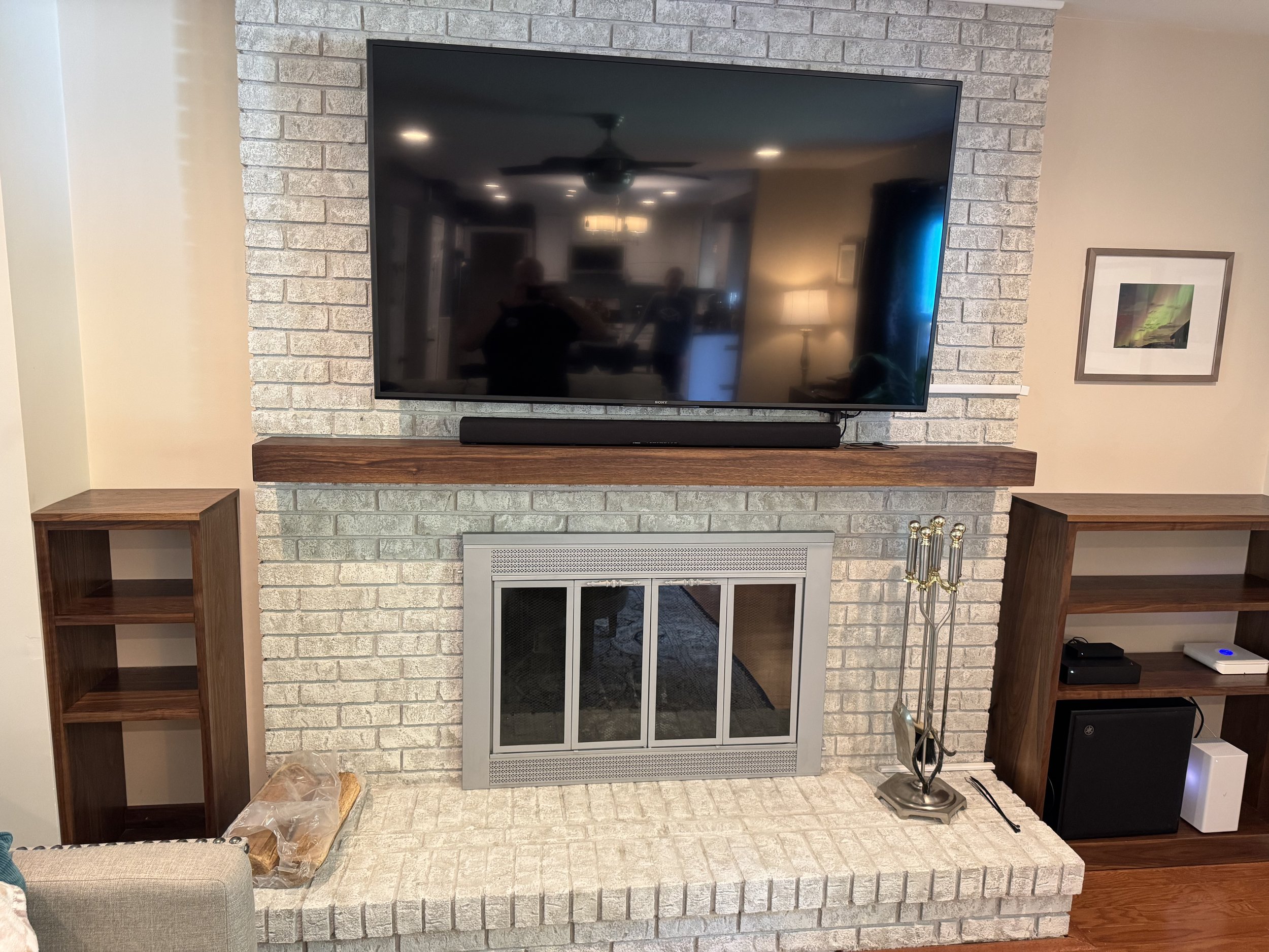 Living room with a brick fireplace and a flat screen TV mounted on the wall above a wooden mantel. On the right, there is a bookshelf with electronic devices and a framed picture on the wall. To the left, there is a small side table with stacks of lo