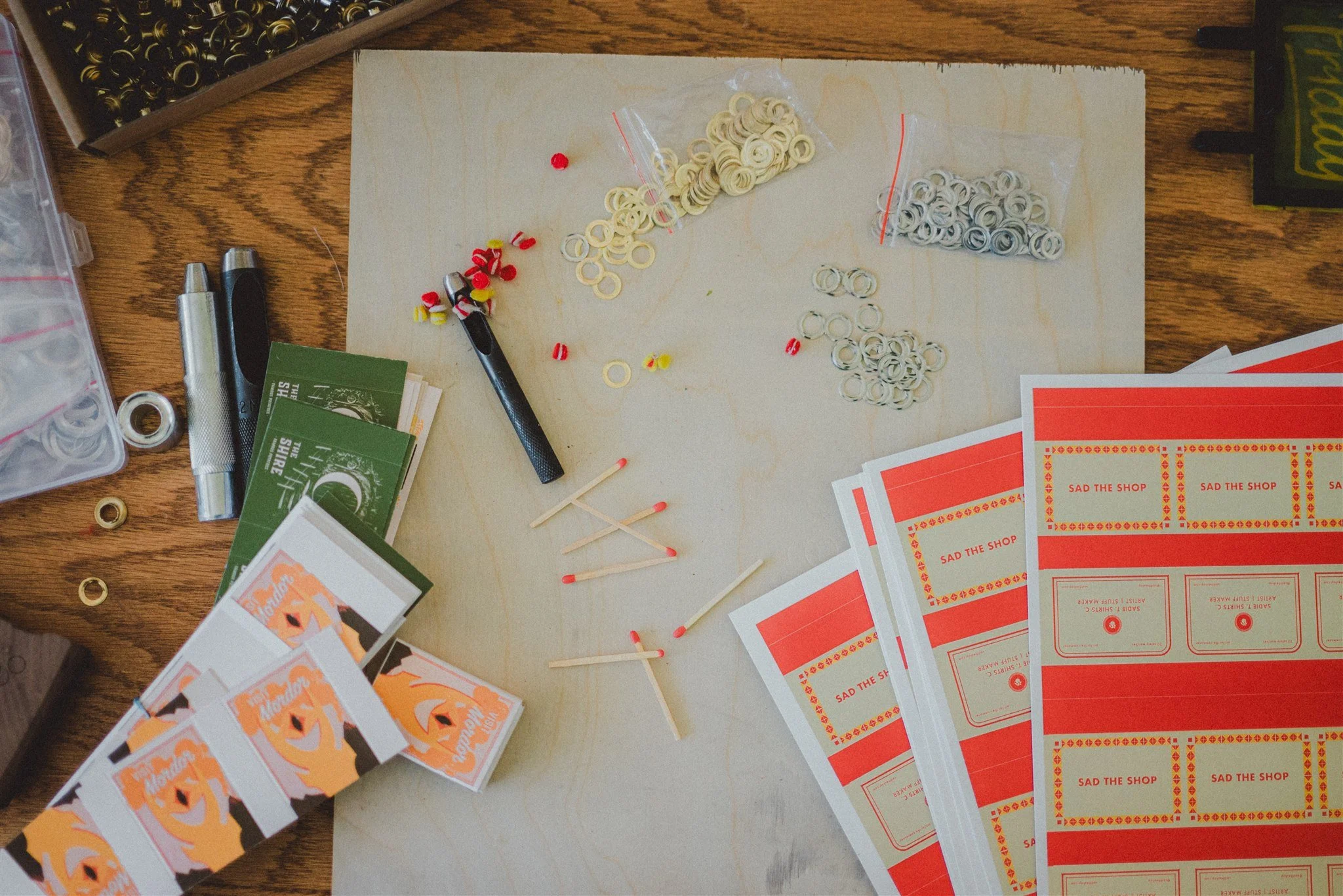 A workspace with small rubber bands, matches, red stickers, and printed labels labeled 'SAD THE SHOP,' along with packaging materials and a box of rubber bands.