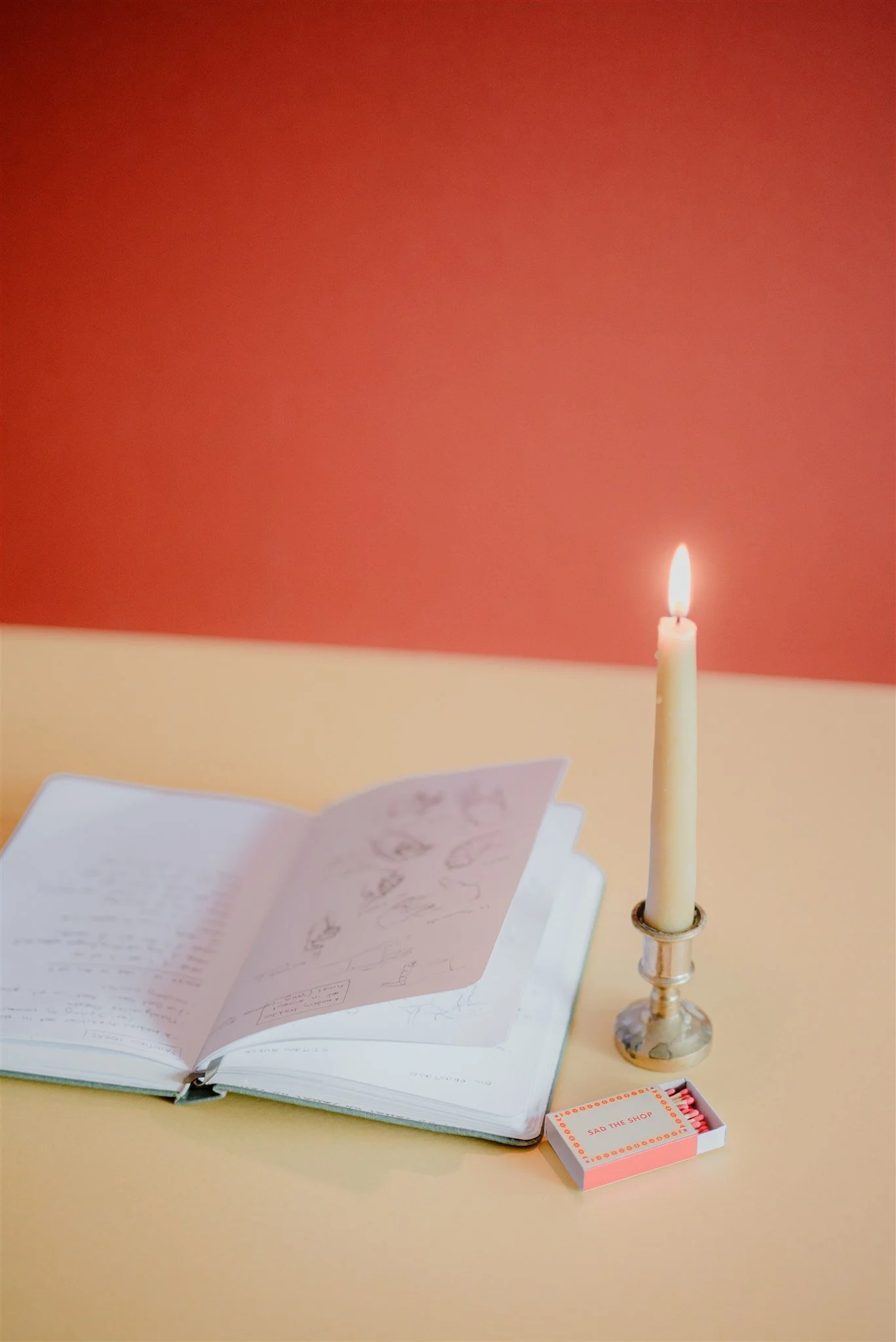 Open sketchbook on yellow table next to a lit candle in a candlestick and a box of pink matches with a label that reads 'Sad the Shop'.