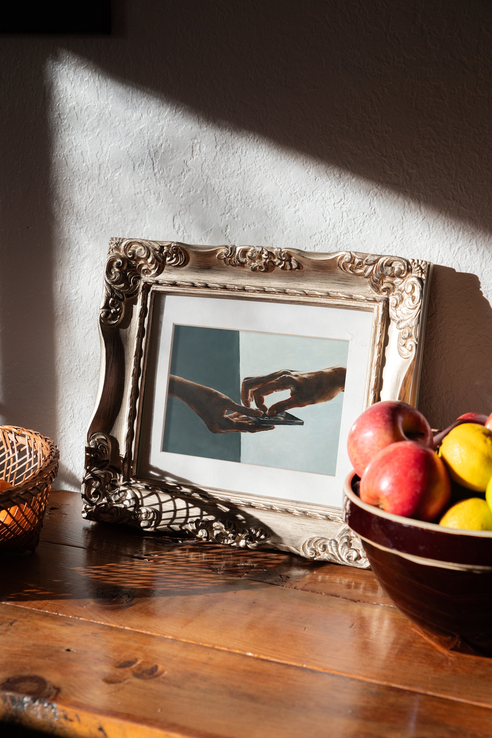 A framed artwork of two hands reaching toward each other, reminiscent of Michelangelo's 'The Creation of Adam,' is placed on a wooden surface next to a bowl of apples and lemons. Original Oil Print from Sadie T. Shirts C., Sad the Shop.