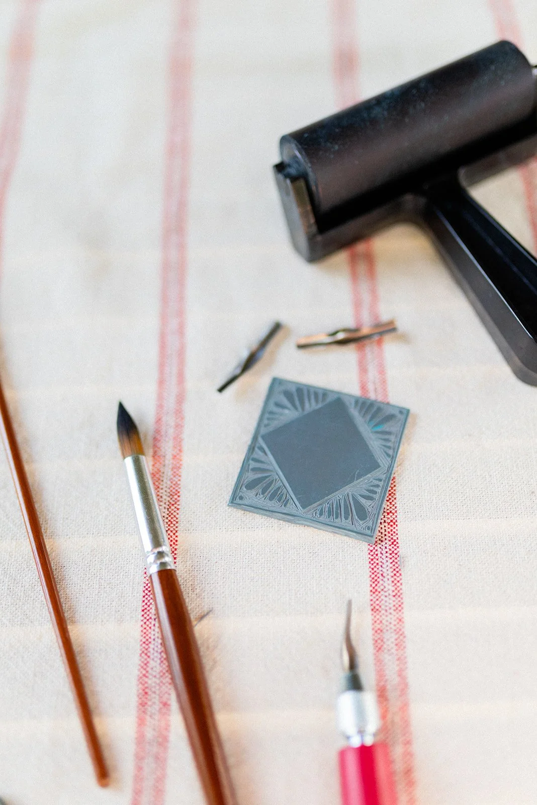 A linoleum stamp, two small engraving tools, a paintbrush, and a pink-handled tattoo needle lie on a striped cloth.