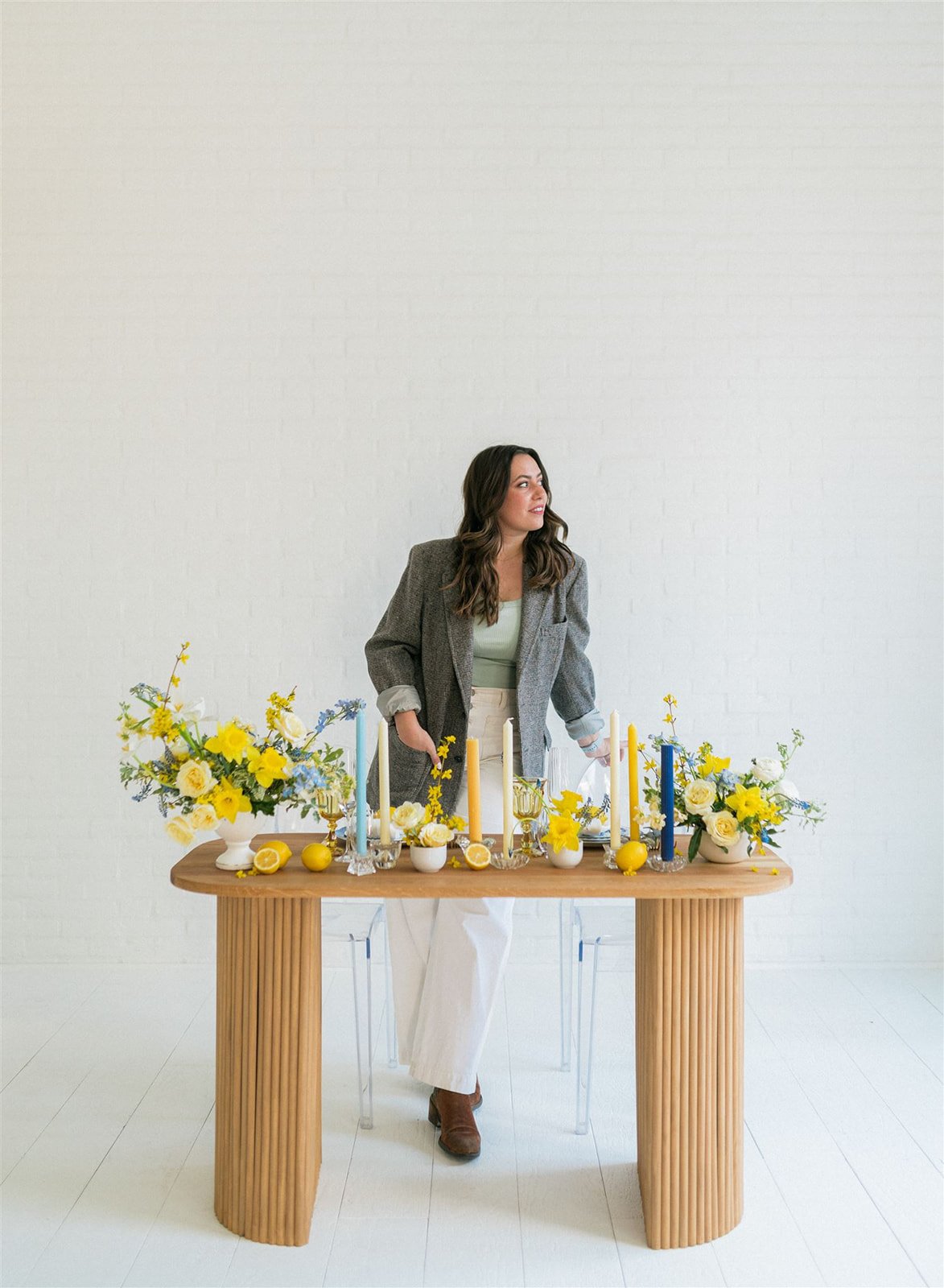 Sadie stands behind a wooden table decorated with yellow and white flowers, lemons, and colorful candles, in a minimalist room with white walls.
