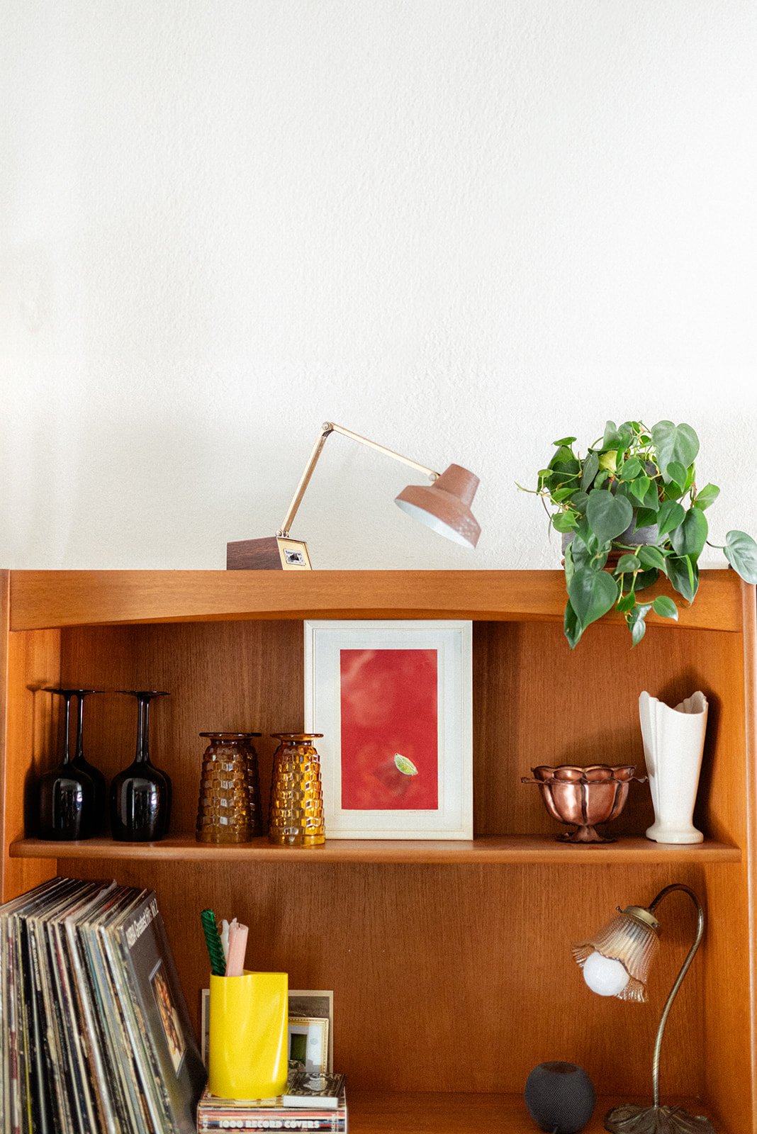A wooden bookshelf filled with decorative items, including a photo frame, a green potted plant, vintage glasses, vases, a small picture, and a desk lamp. Original Lime Painting by Sadie T. Shirts C., Sad the Shop.