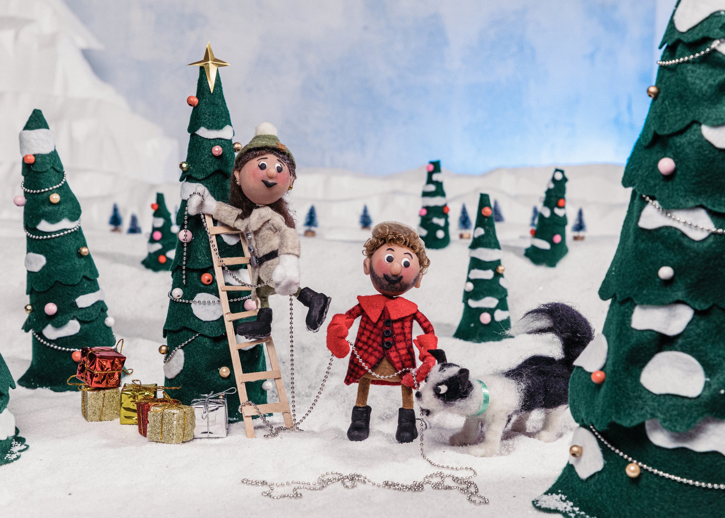 Felt characters in a snowy Christmas scene, with trees decorated with ornaments, presents, and a backdrop of blue sky. Original Stop Motion Artwork from Sadie T. Shirts C., Sad the Shop.