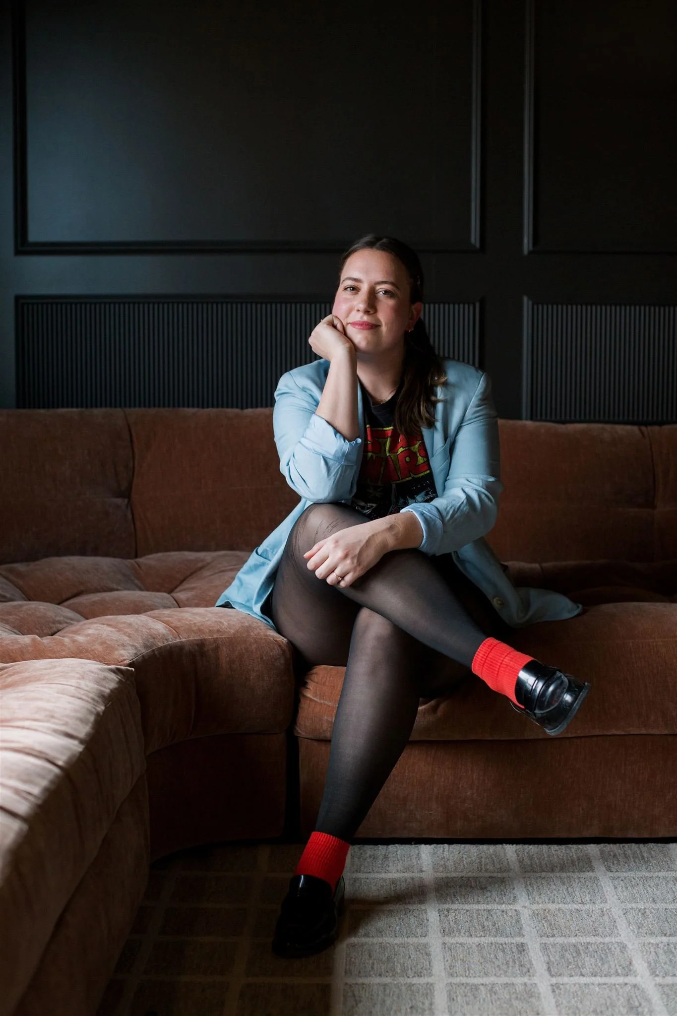 Sadie sitting on a brown couch in a dimly lit room, wearing black tights, a Star Wars t-shirt, a blue blazer, and black shoes with red socks, with her legs crossed and resting her chin on her hand, smiling