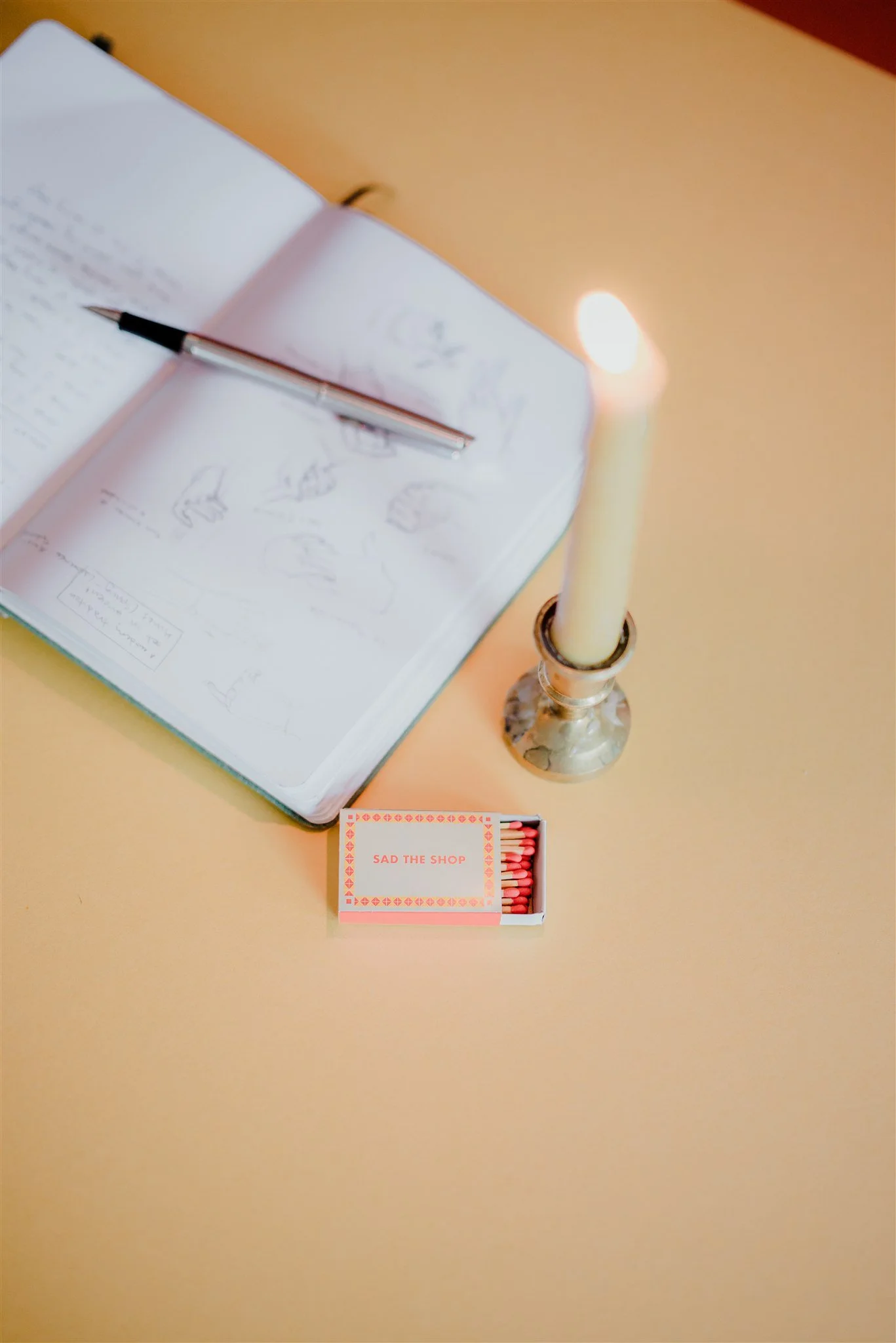 Open sketchbook with drawings and a pen, a lit candle in a silver holder, and a box of matches labeled 'SAD THE SHOP' on a yellow surface.