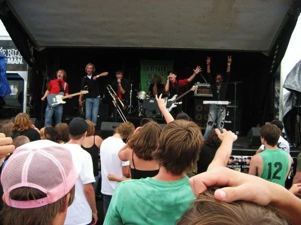 A band performs on an outdoor stage during warped tour. The audience, mostly children and teenagers, watches and some raise their hands.
