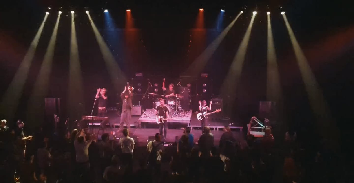 A band performs on stage at a concert with colorful stage lighting and an audience in front.