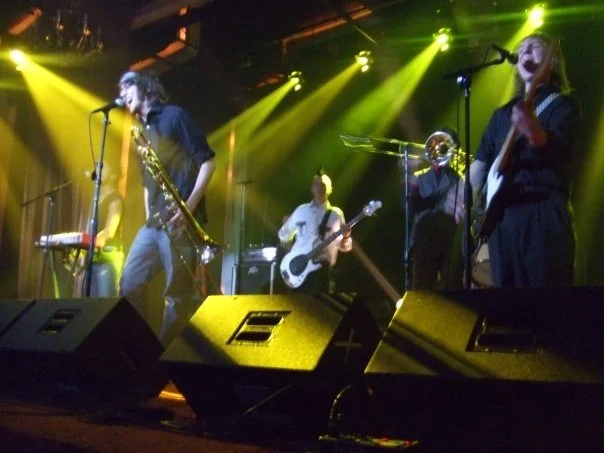 Band of musicians performing on stage with yellow and green stage lights, including a trombonist, a guitarist, and a bassist, in a concert setting.