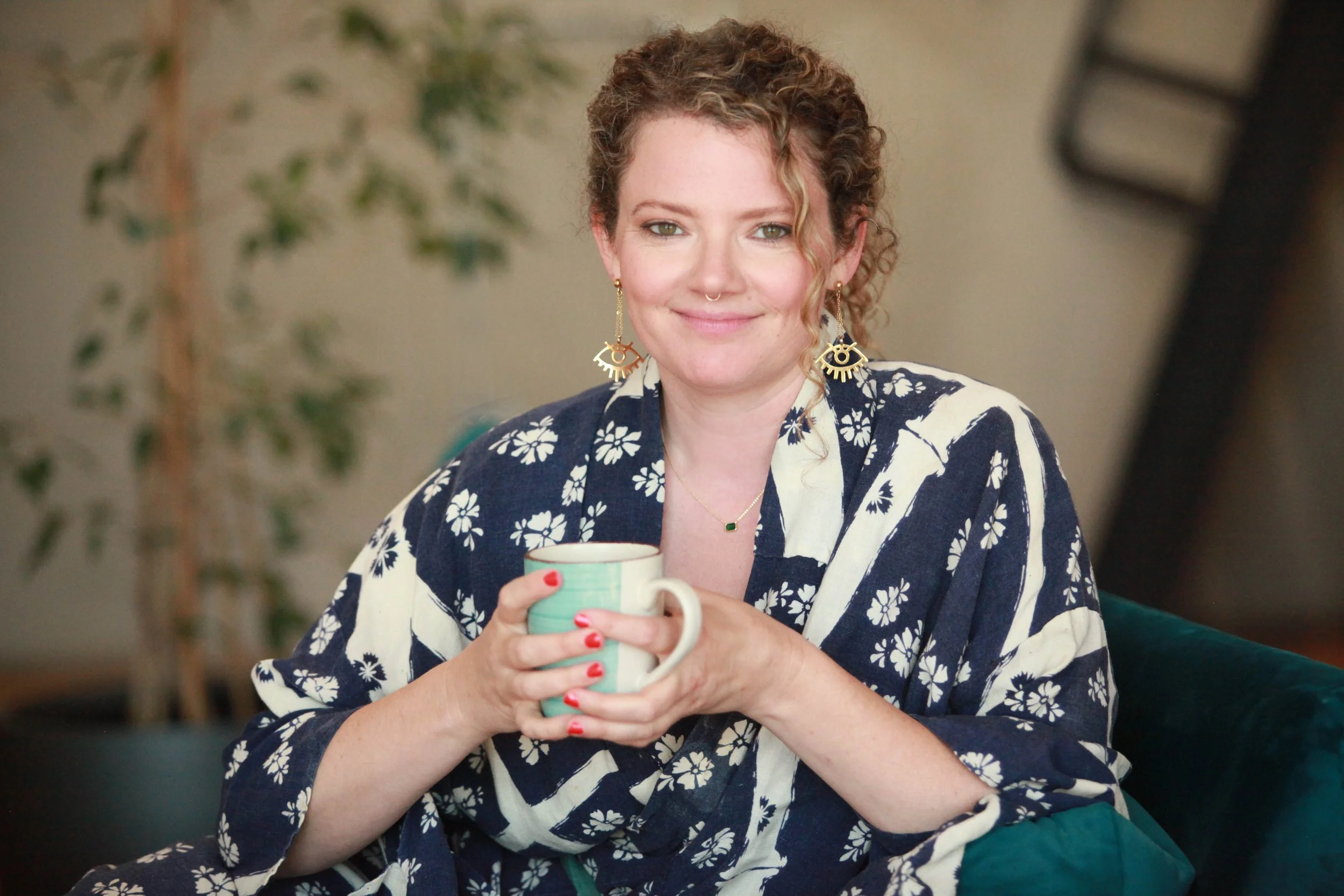 Emily Webb seated in a floral-patterned top, holding a green mug and smiling.