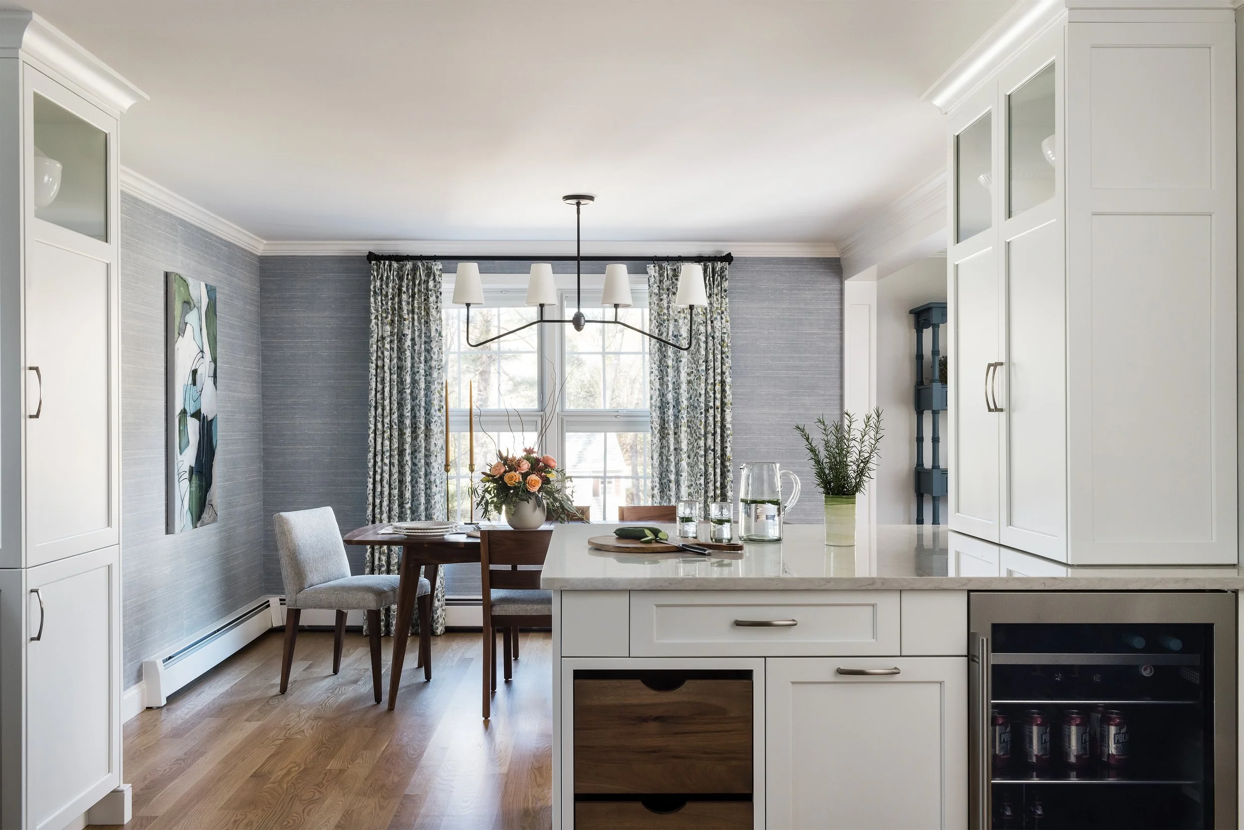 Modern kitchen and dining area with white cabinetry, a marble countertop, a small wine fridge, a dining table with four chairs, floral centerpiece, window with patterned curtains, and hardwood floors.