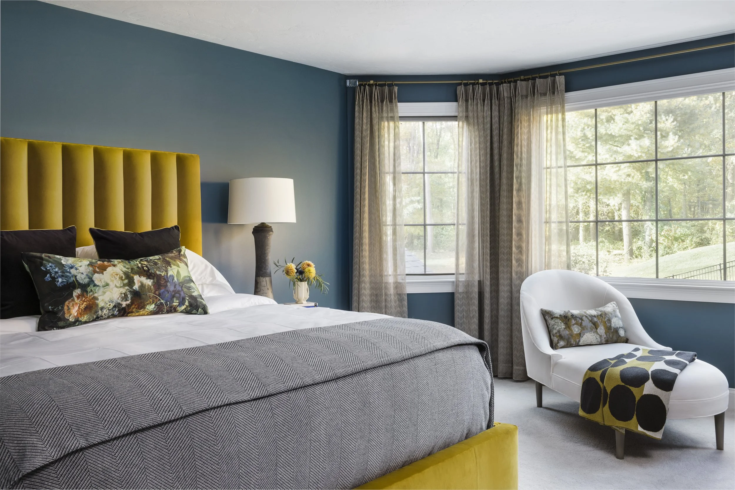 A bedroom with a large bed having a yellow headboard, floral and black pillows, gray bedspread, and white sheets. Next to the bed is a nightstand with a white lamp and a vase with flowers. The room has large windows with sheer and patterned curtains,