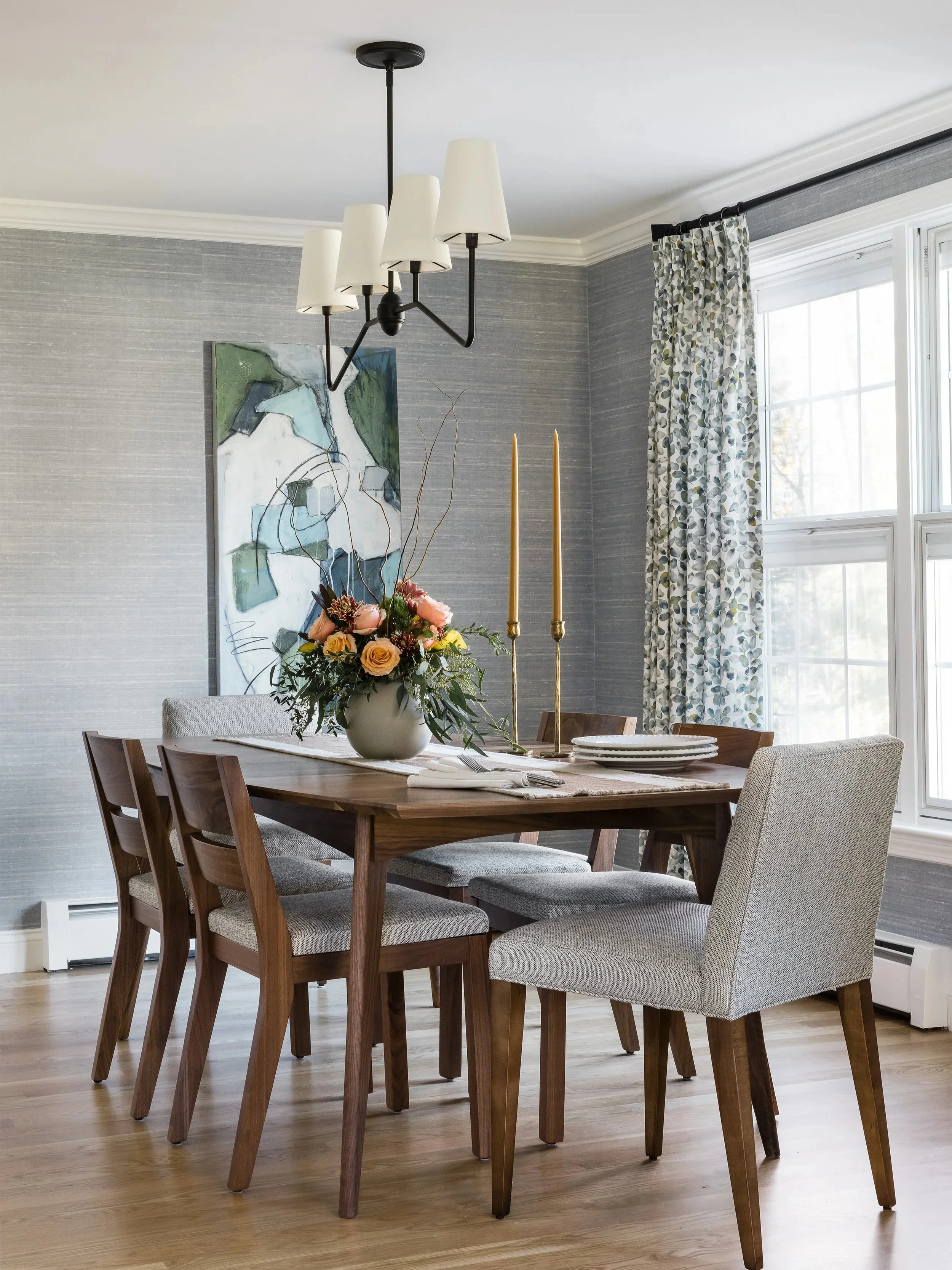 A dining room with a wooden table and six chairs, a floral centerpiece, two tall gold candlesticks, a ceiling light fixture, large windows with floral curtains, and artwork on textured gray walls.