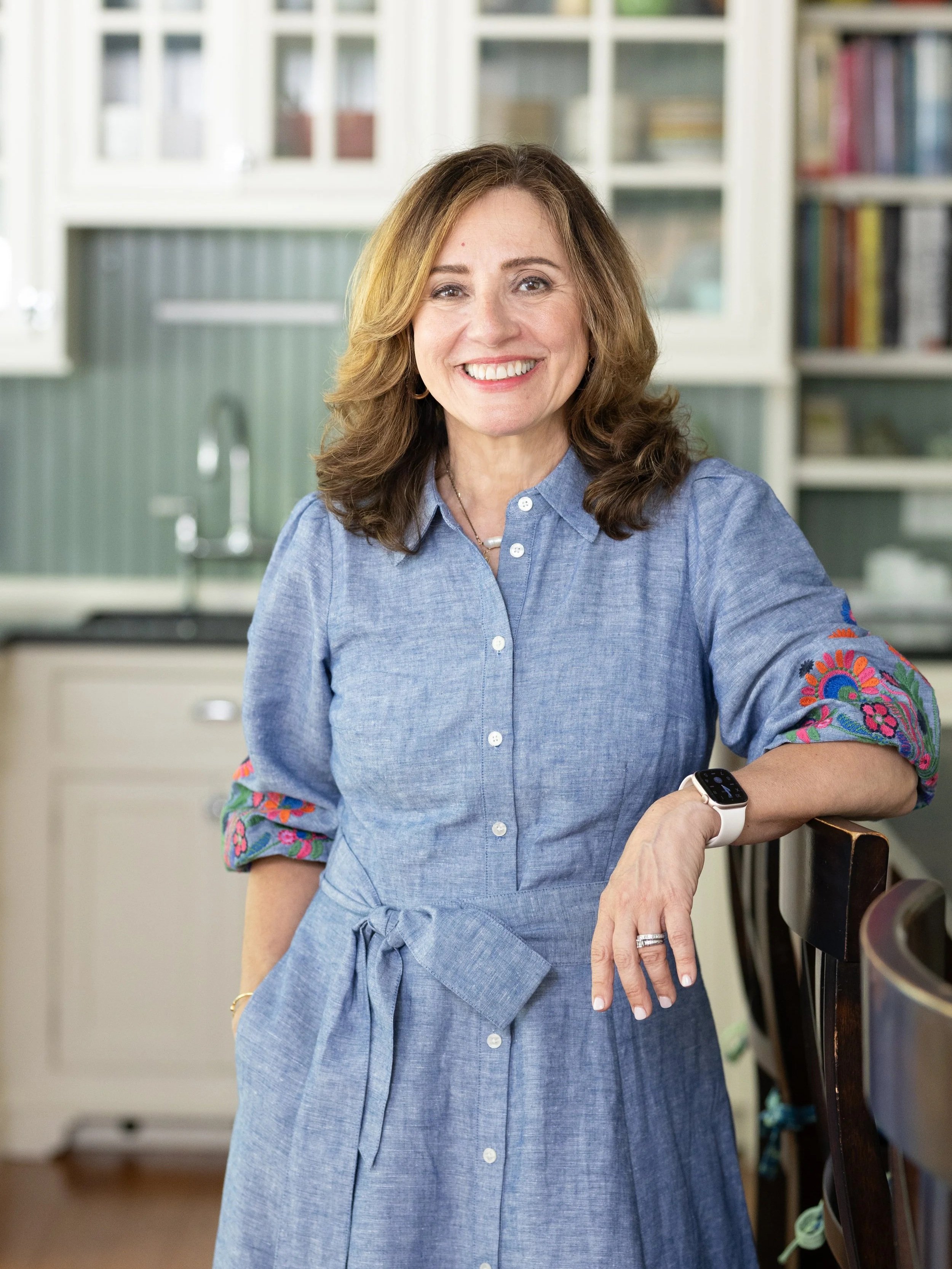A woman in a denim dress with embroidered sleeves, smiling and standing in a kitchen.