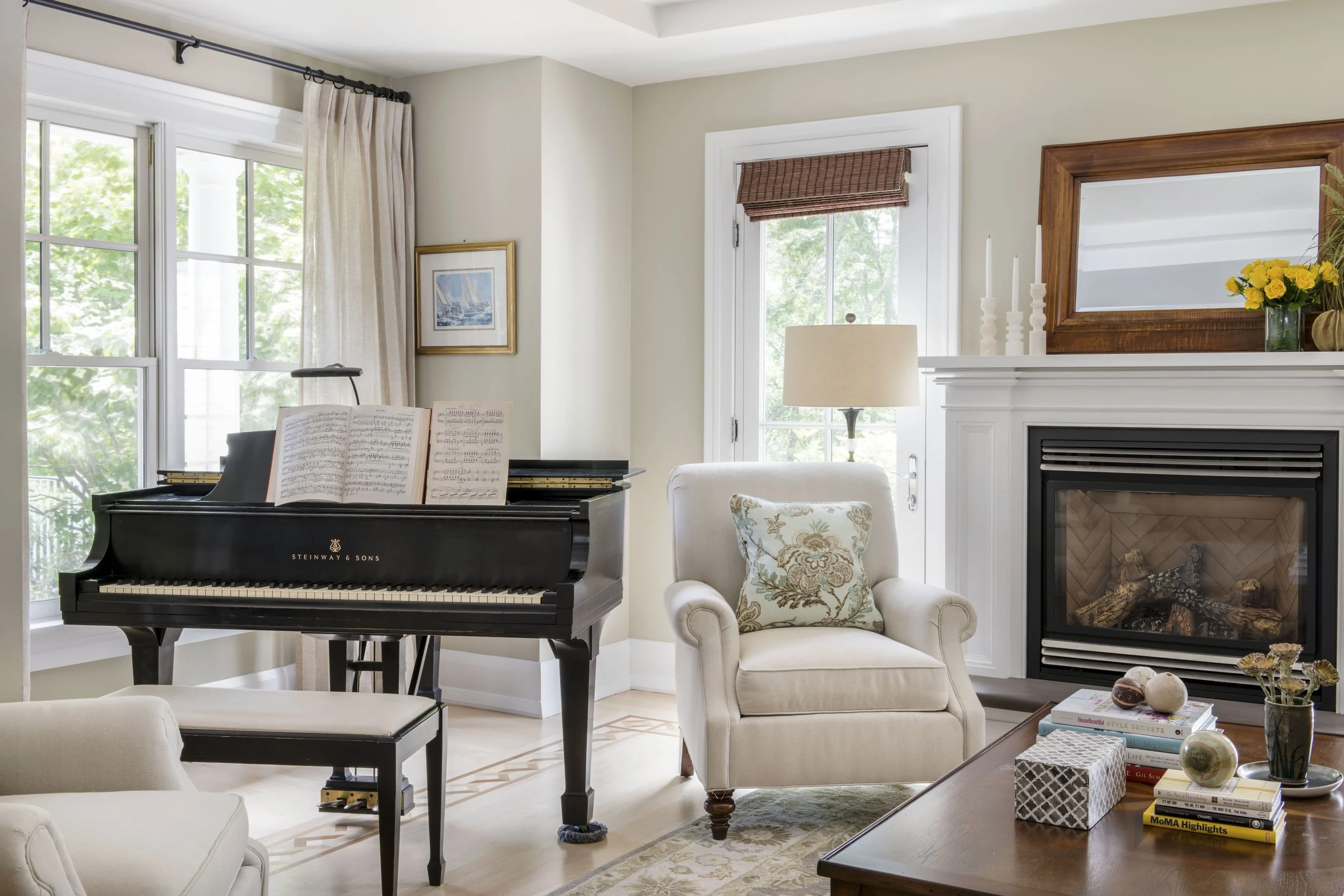 Living room with Steinway & Sons grand piano, white armchair with floral pillow, fireplace with mirror and yellow flowers, side table with books and decorative items, window with curtains, artwork on walls, and a door with a brown window shade.