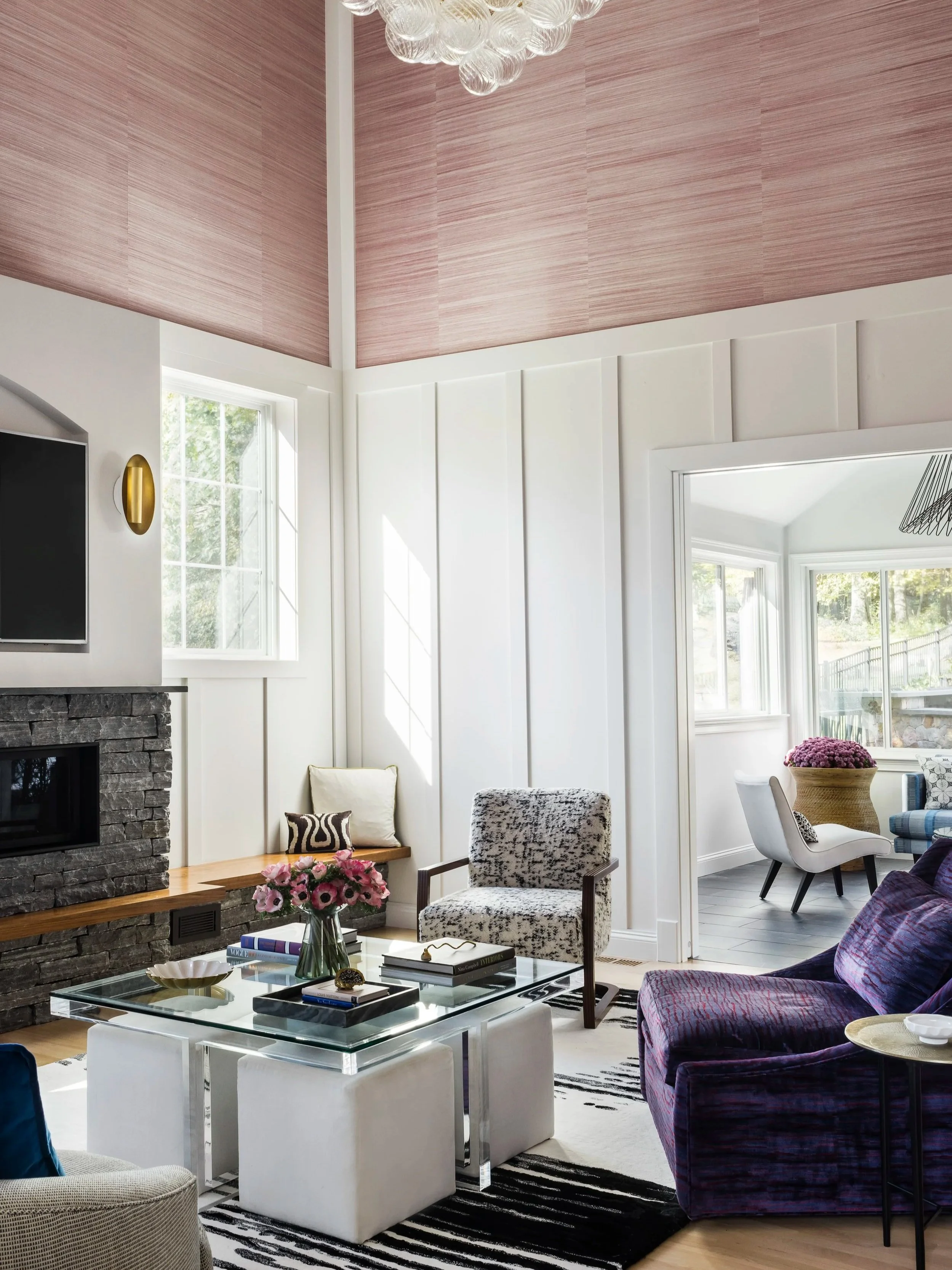 Living room with black stone fireplace, white walls, pink ceiling, and large windows; furniture includes a glass coffee table with books and a flower vase, a purple sofa, patterned armchair, and white ottomans.