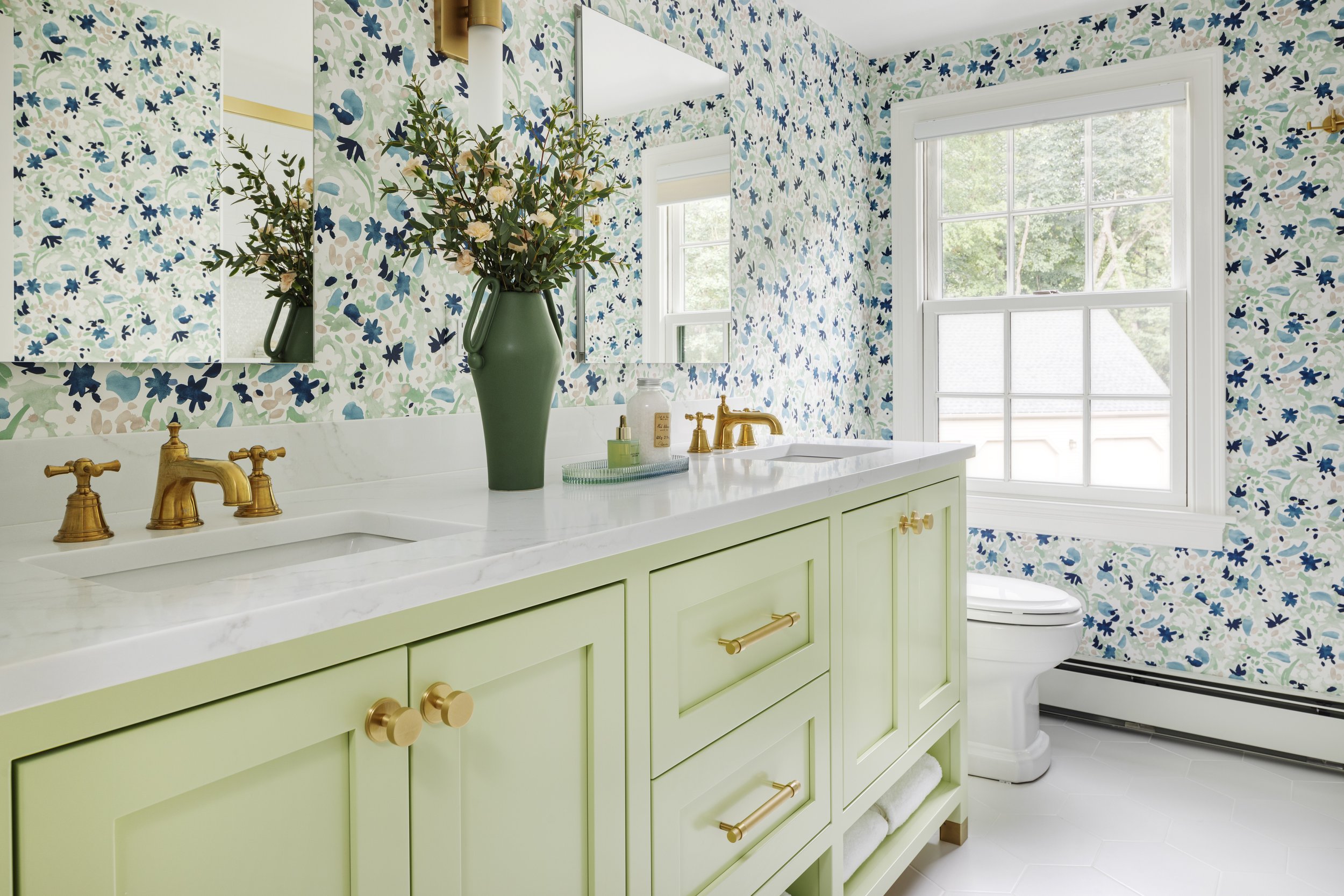 A bathroom with a green vanity, gold fixtures, a large mirror, a patterned wallpaper with blue and green floral designs, a large window, and a vase with flowers on the counter.