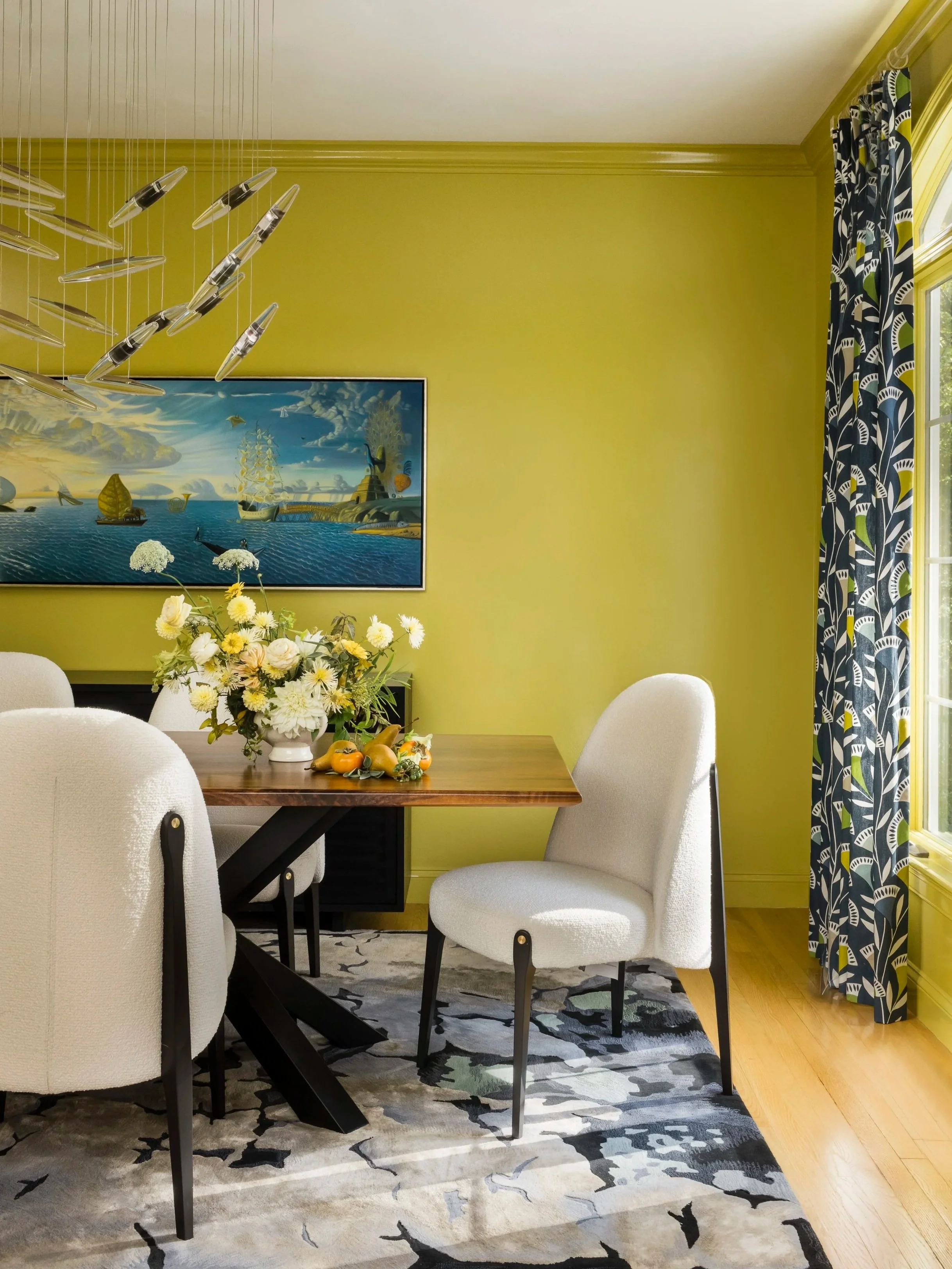 Bright yellow wall dining room with a wooden table, four white upholstered chairs, a large floral centerpiece, a painting of a seascape on the wall, a window with blue patterned curtains, and a black and white abstract rug on a wooden floor.