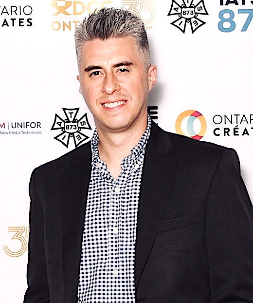 A young man with short gray hair, wearing a black blazer over a checkered shirt, standing in front of a backdrop with multiple logos and text.