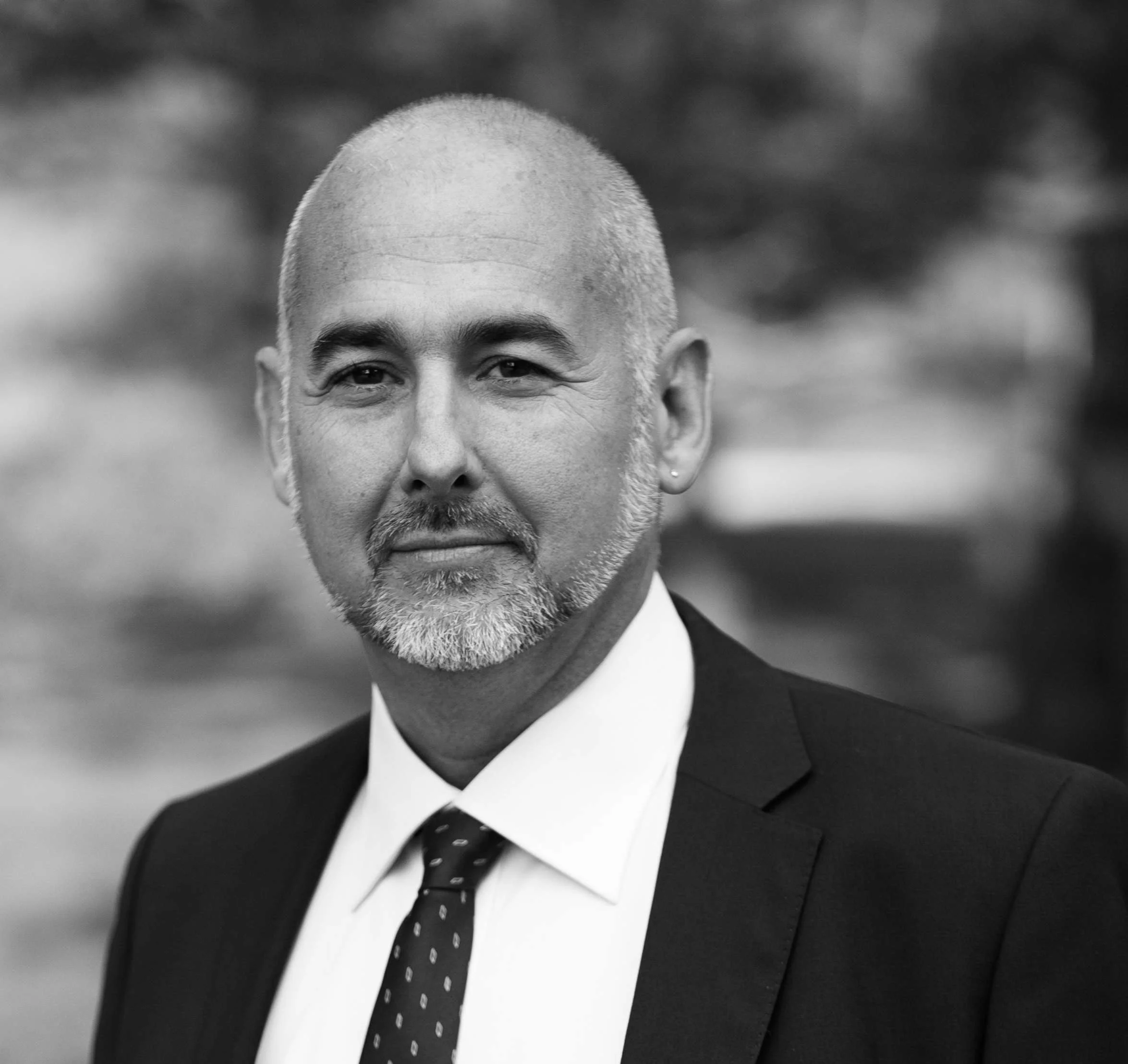 Black and white professional headshot of a middle-aged man in a suit and tie, with a buzz cut and a short beard, smiling slightly.