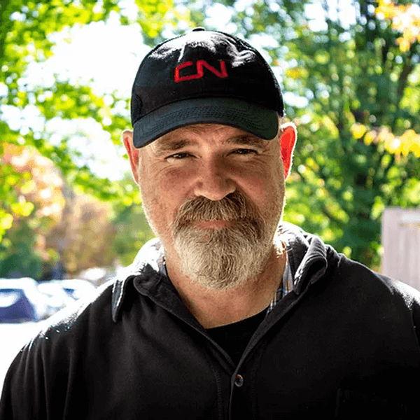 A man with a beard wearing a black cap with red text, a black jacket, and a checkered shirt outdoors with green trees and parked cars in the background.
