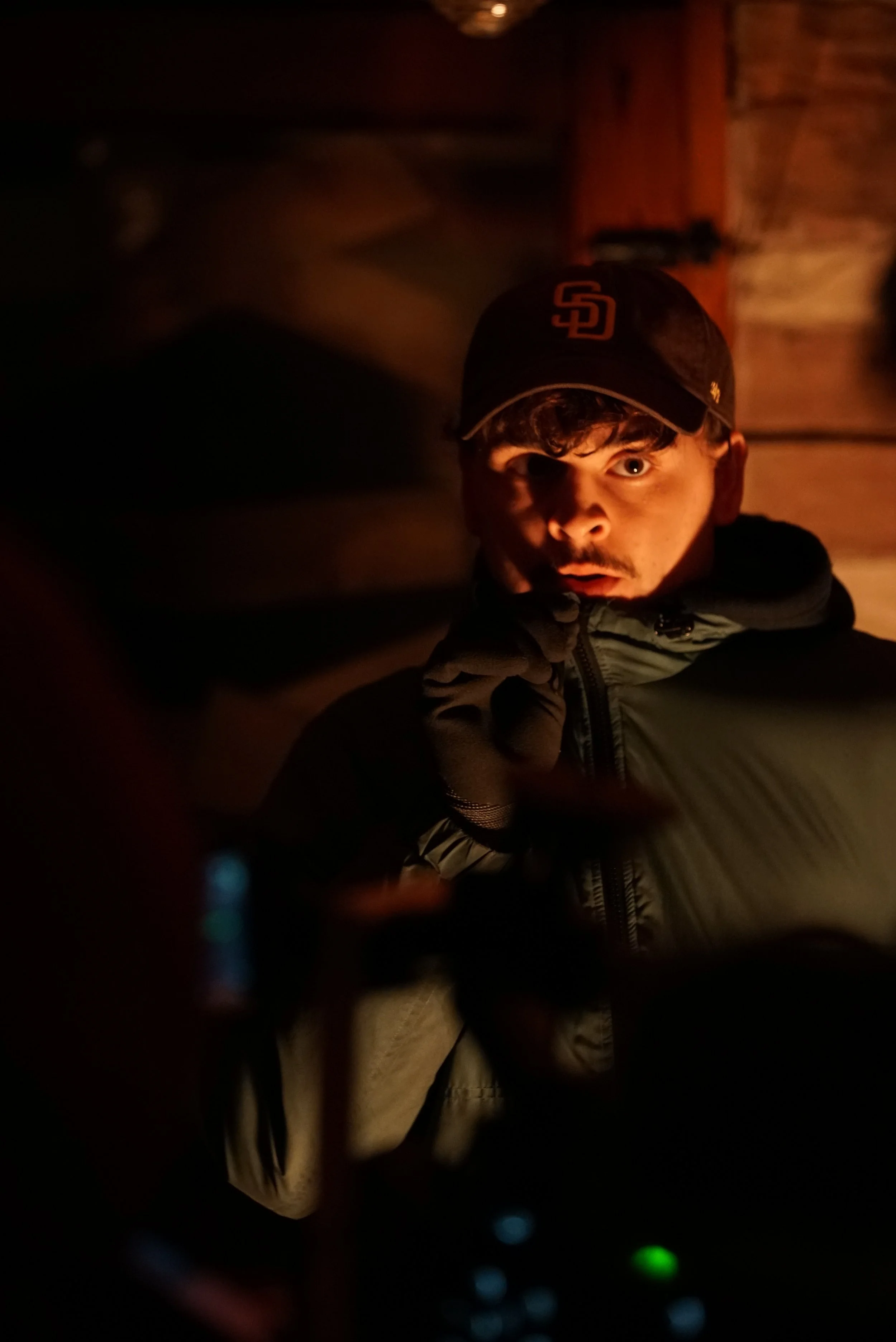 Young man in a dark room with minimal lighting, wearing a black cap with an 'SD' logo and a gray jacket, holding a phone to his ear, with a surprised or intrigued expression.