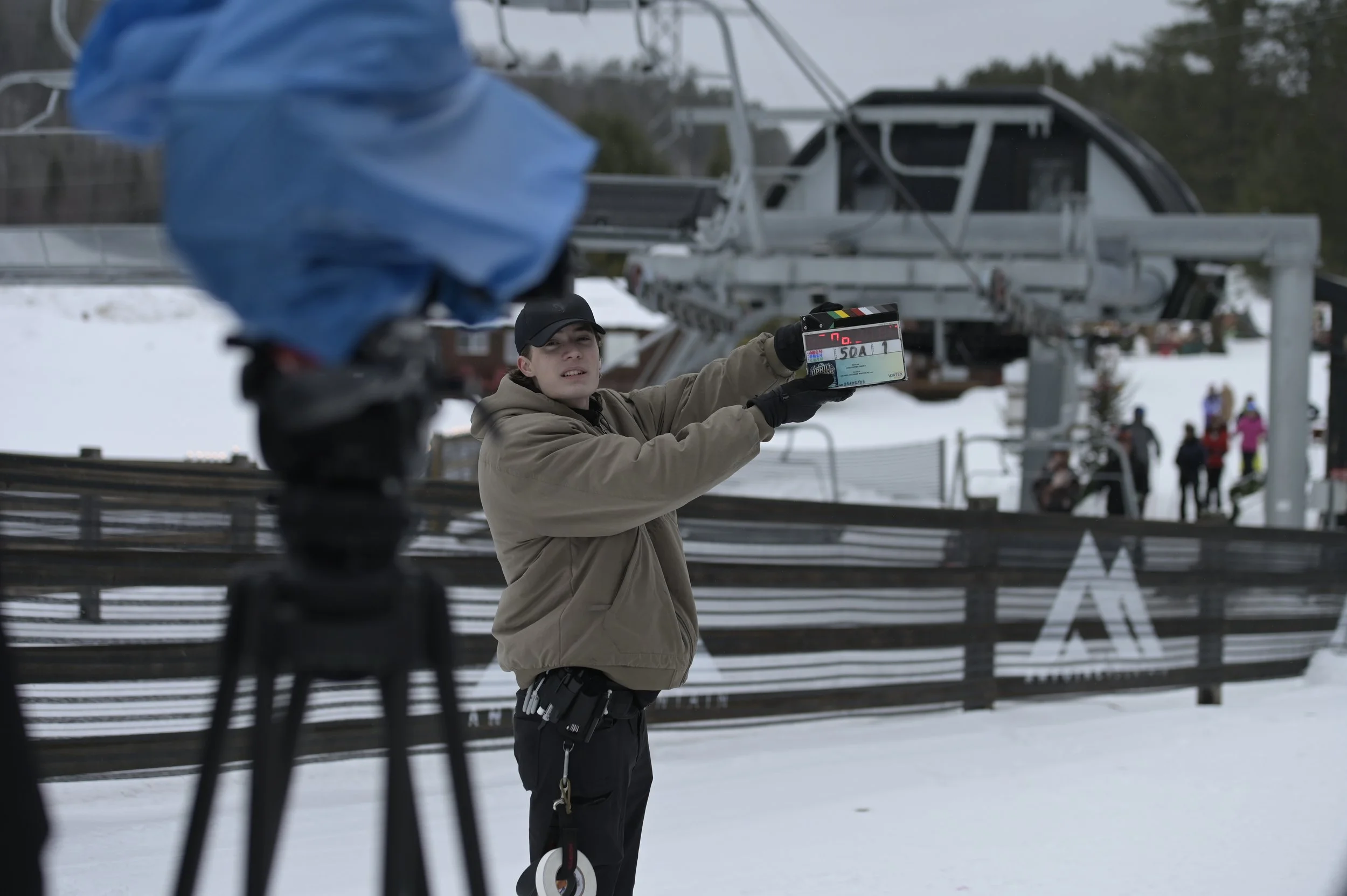 A film crew member holding a clapperboard outdoors in a snowy ski resort with ski lifts and skiers in the background.