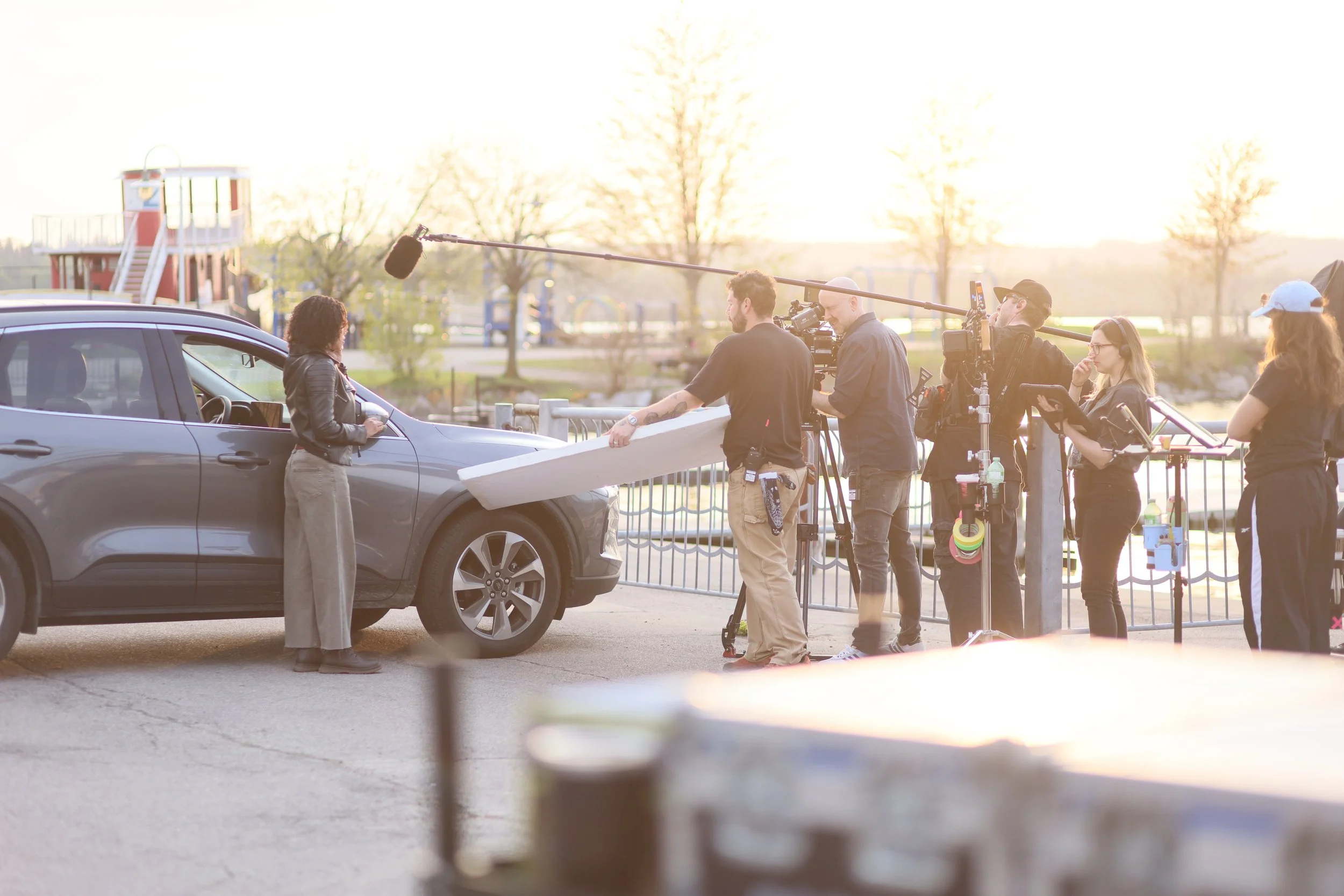 Film crew setting up equipment to film a scene with a woman standing beside a gray car at a park during sunset.