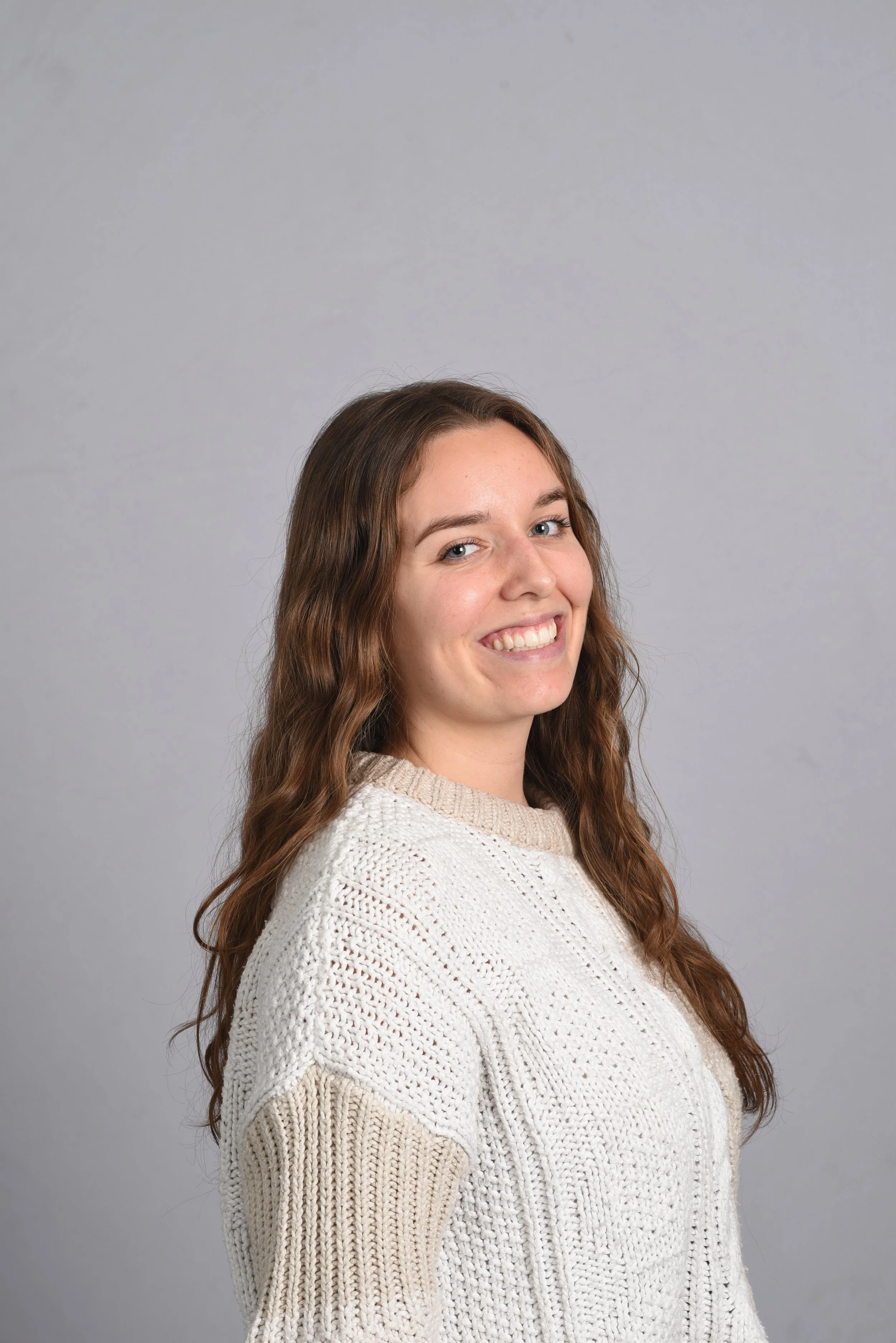A young woman with long wavy brown hair smiling and looking at the camera, wearing a cream knitted sweater, standing against a plain gray background.