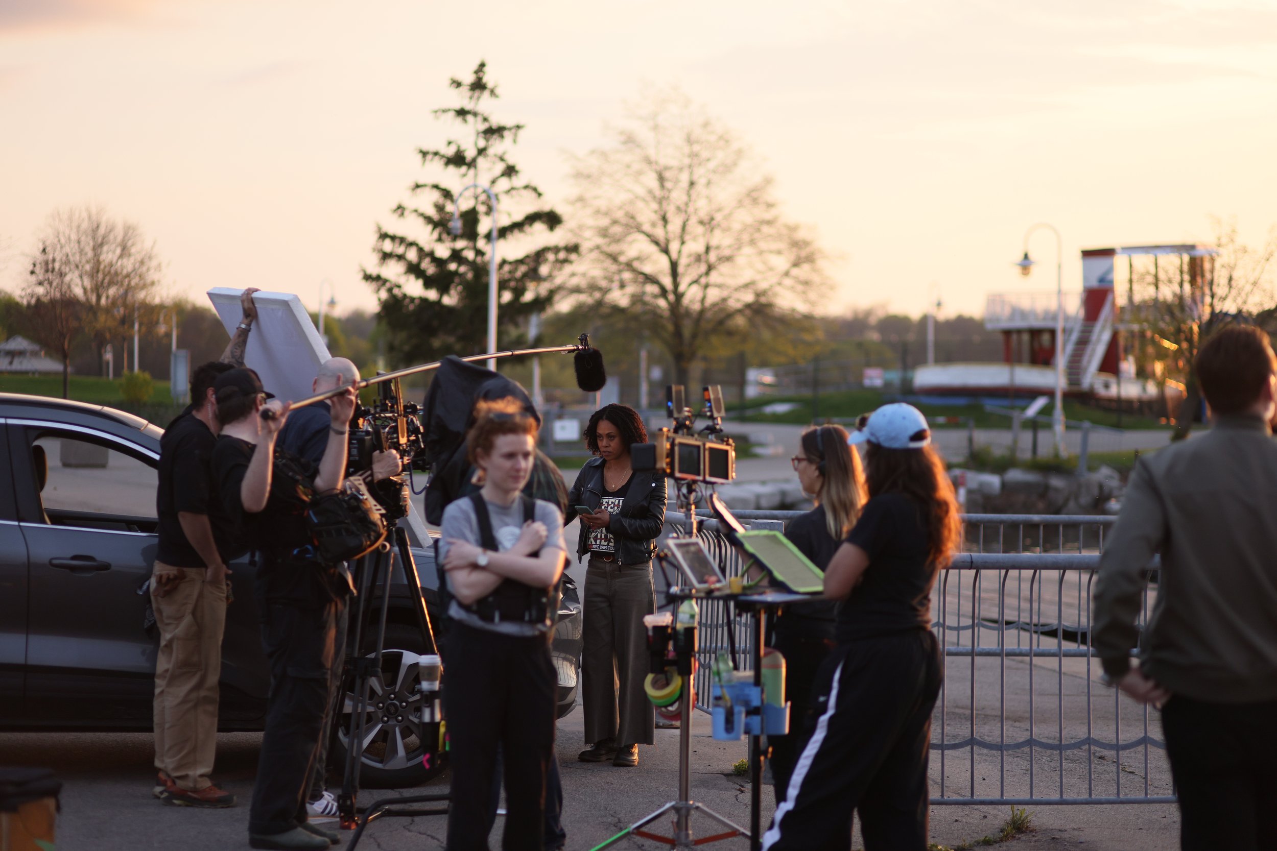 Filmmakers filming outdoors at sunset, with cameras, microphones, and crew members near a black car and a barrier, trees, and a small building in the background.