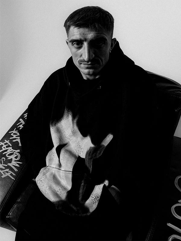 Black and white photo of a man sitting in a chair, wearing a hoodie with a graphic print, with graffiti on the armrest.