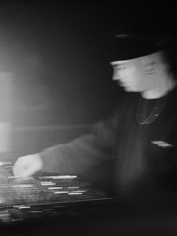 A person wearing a hat and dark clothing is blurred while working with DJ equipment or a mixer, with a light source creating lens flare on the left side.