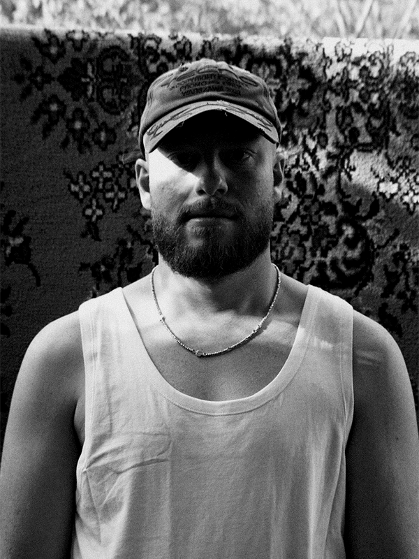 A black and white photo of a bearded man wearing a baseball cap, tank top, and necklace, standing in front of a patterned textile background.